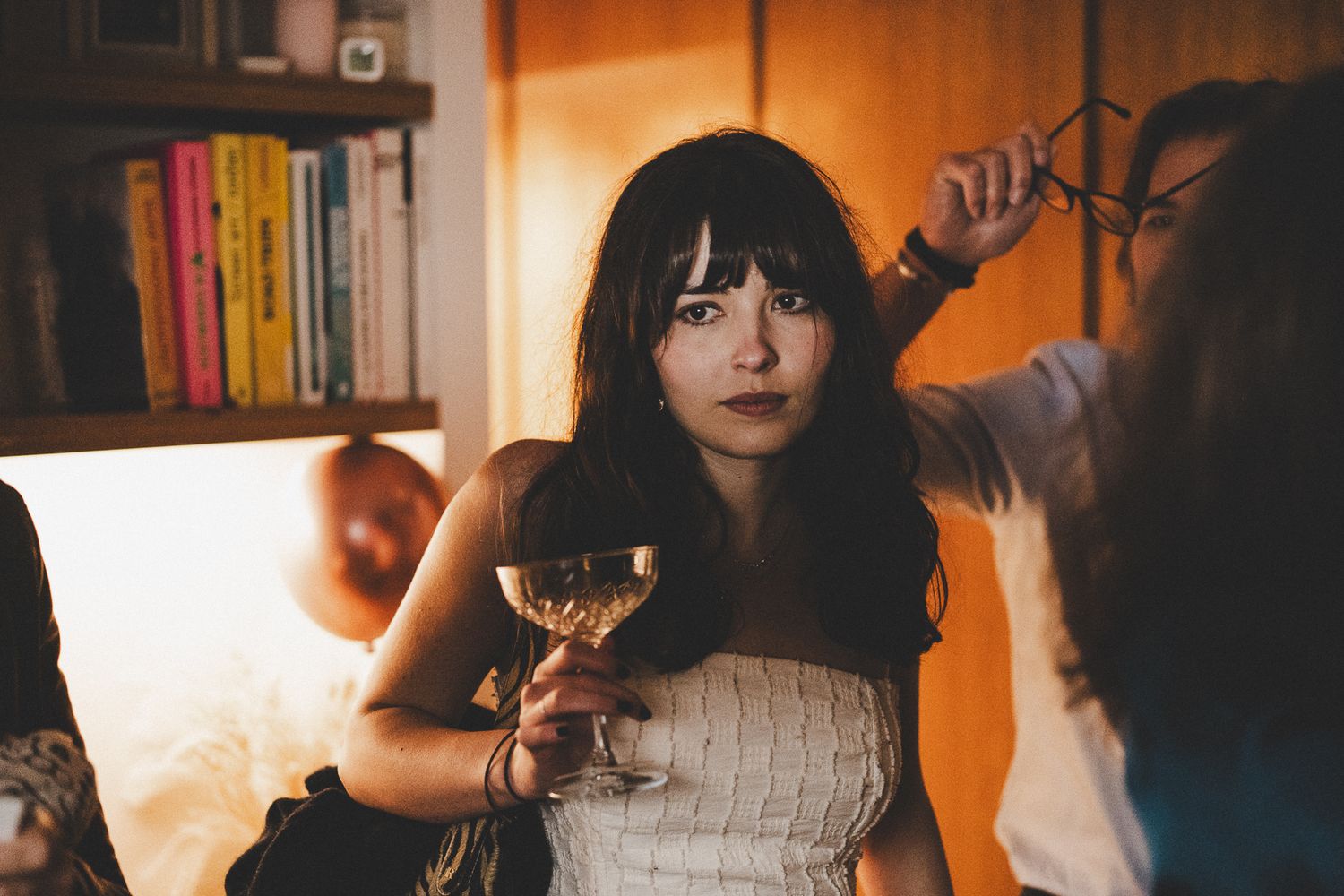 Young woman with dark hair holding a wine glass at an indoor party, looking thoughtful.
