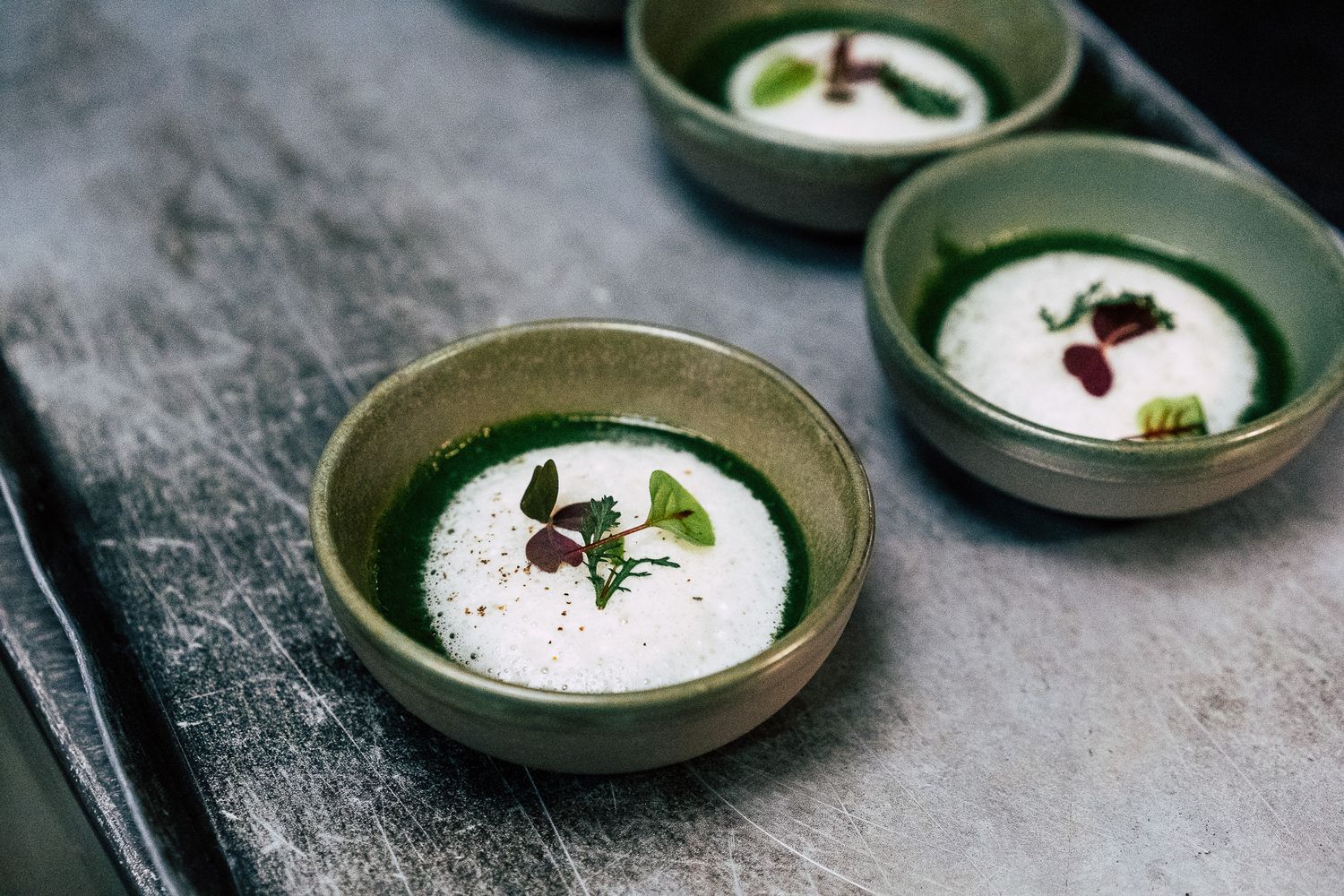Three green ceramic bowls with creamy white soup garnished with fresh herbs on a dark surface.