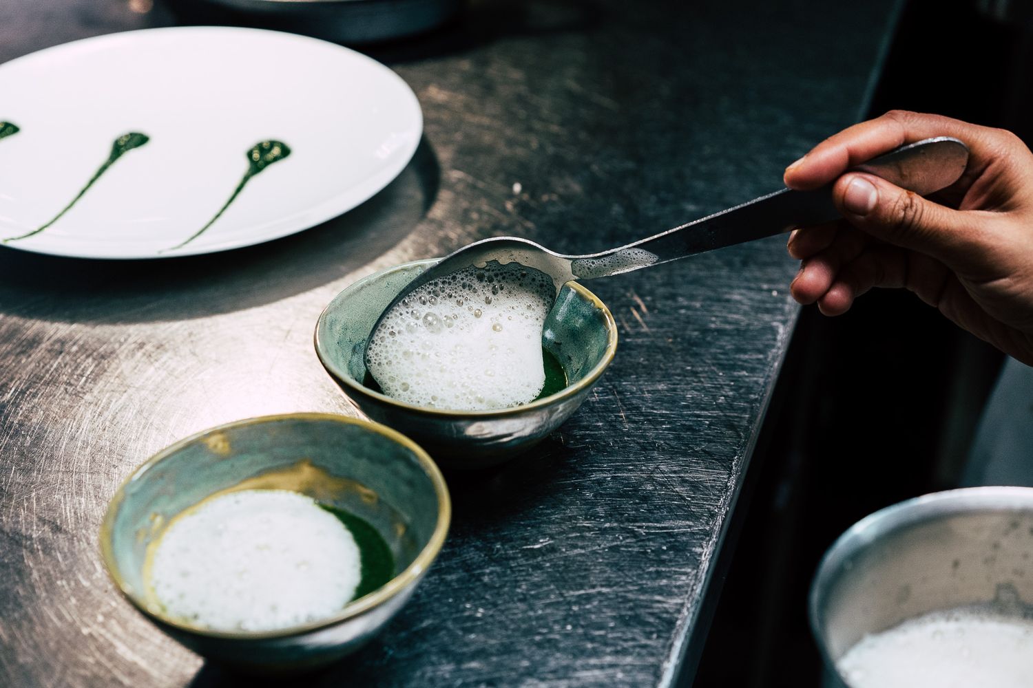 A chef's hand uses tongs to place a round salt or sugar mound into a rustic green ceramic bowl on a dark surface.