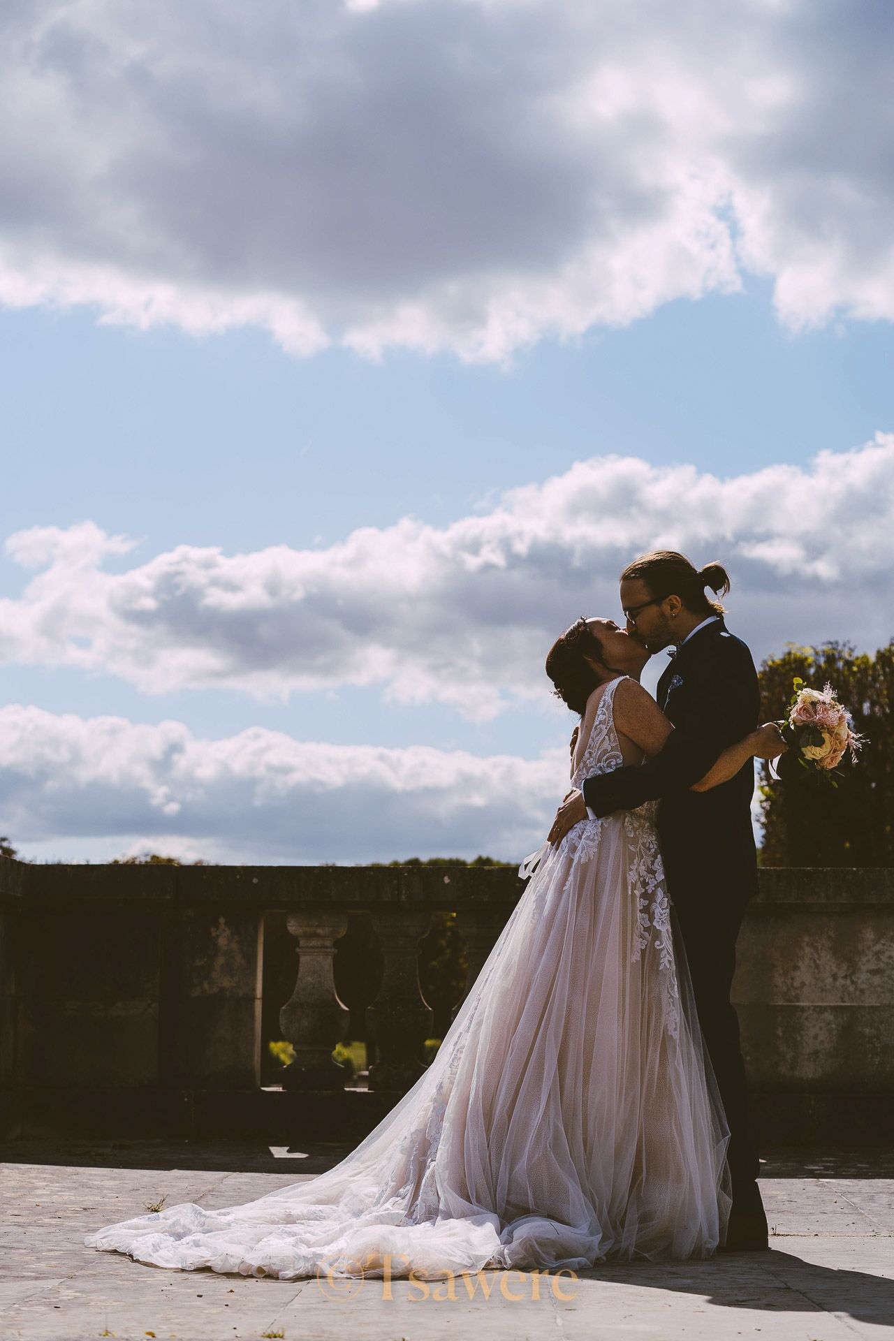Bride and groom sharing romantic moment on terrace under beautiful blue sky with clouds.