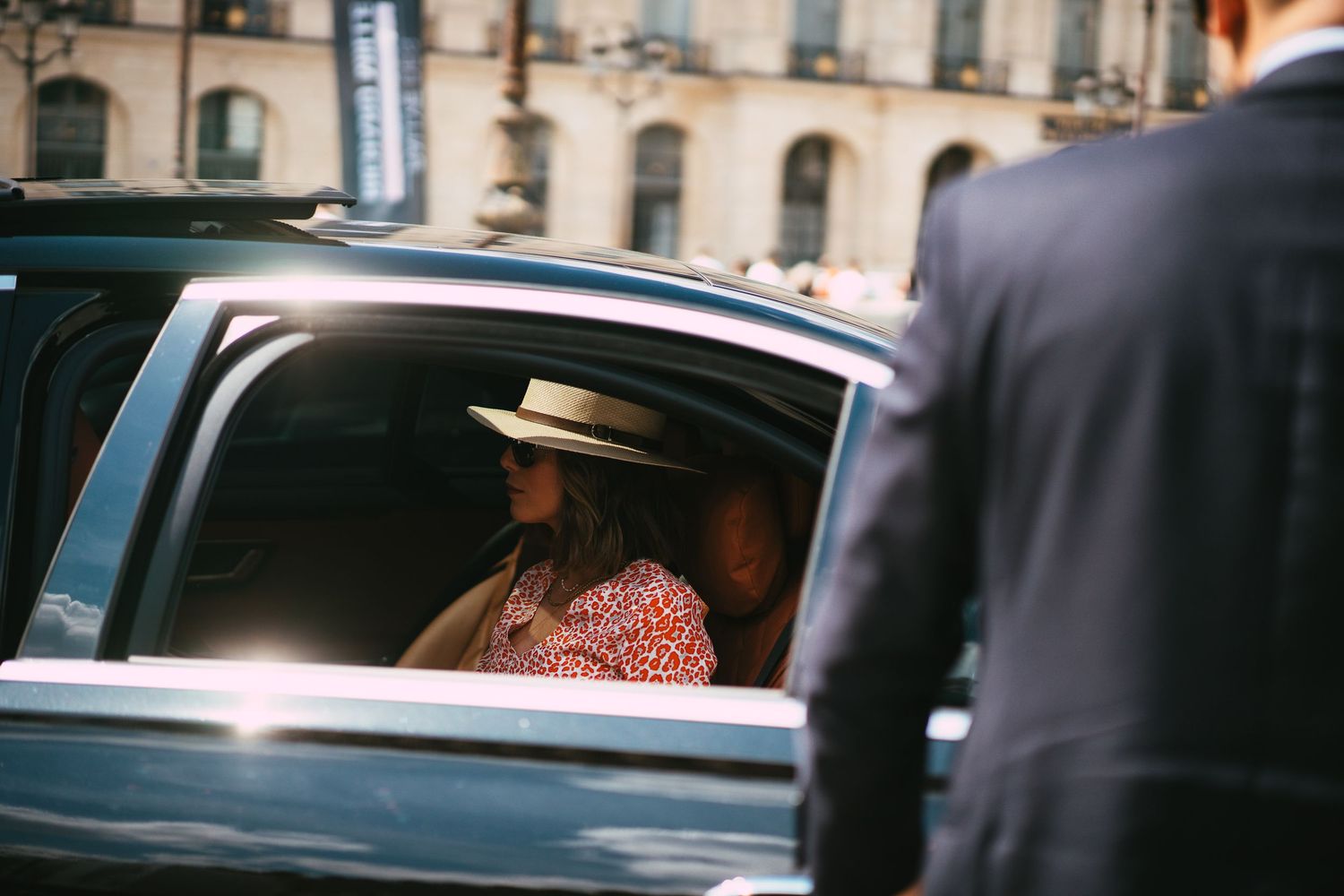 Woman in floral dress and hat seated in luxury car backseat with suited driver standing outside.
