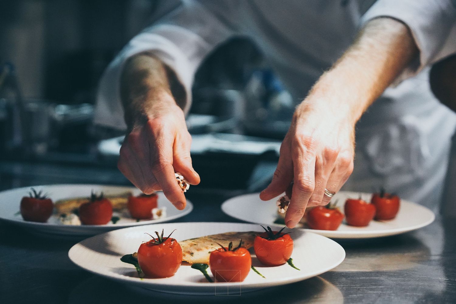 Chef plating cherry tomatoes on white dishes in a professional kitchen.