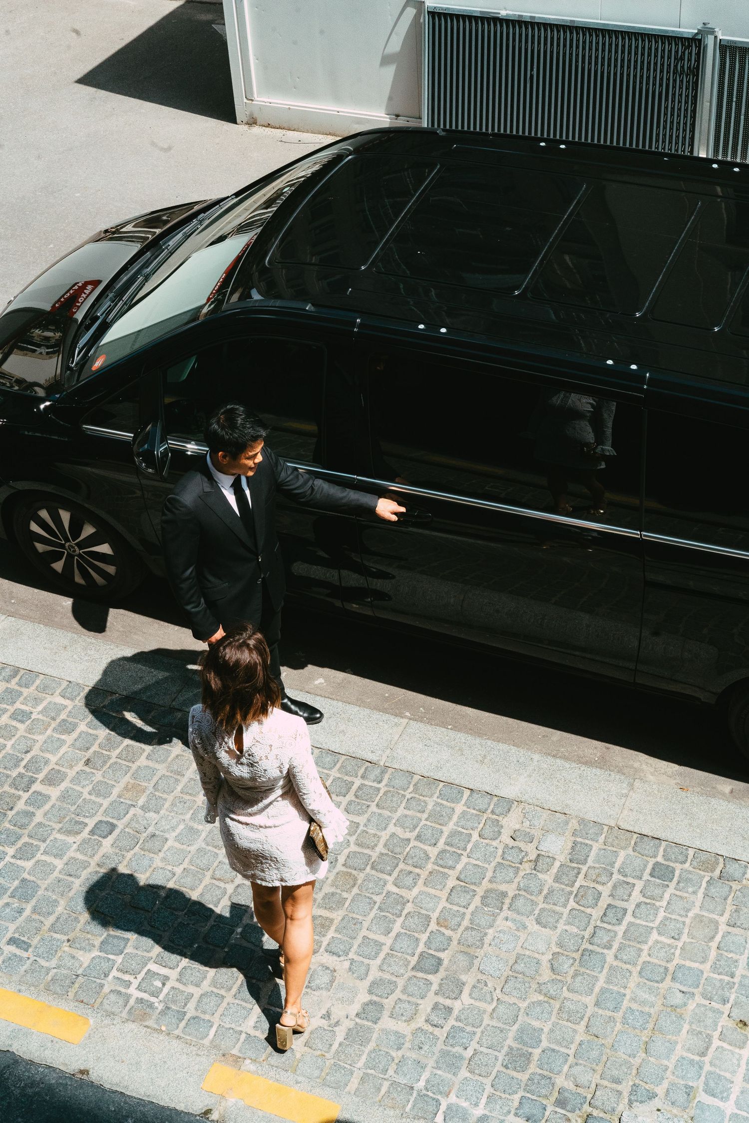 Aerial view of chauffeur opening door of black luxury van for woman on cobblestone street.
