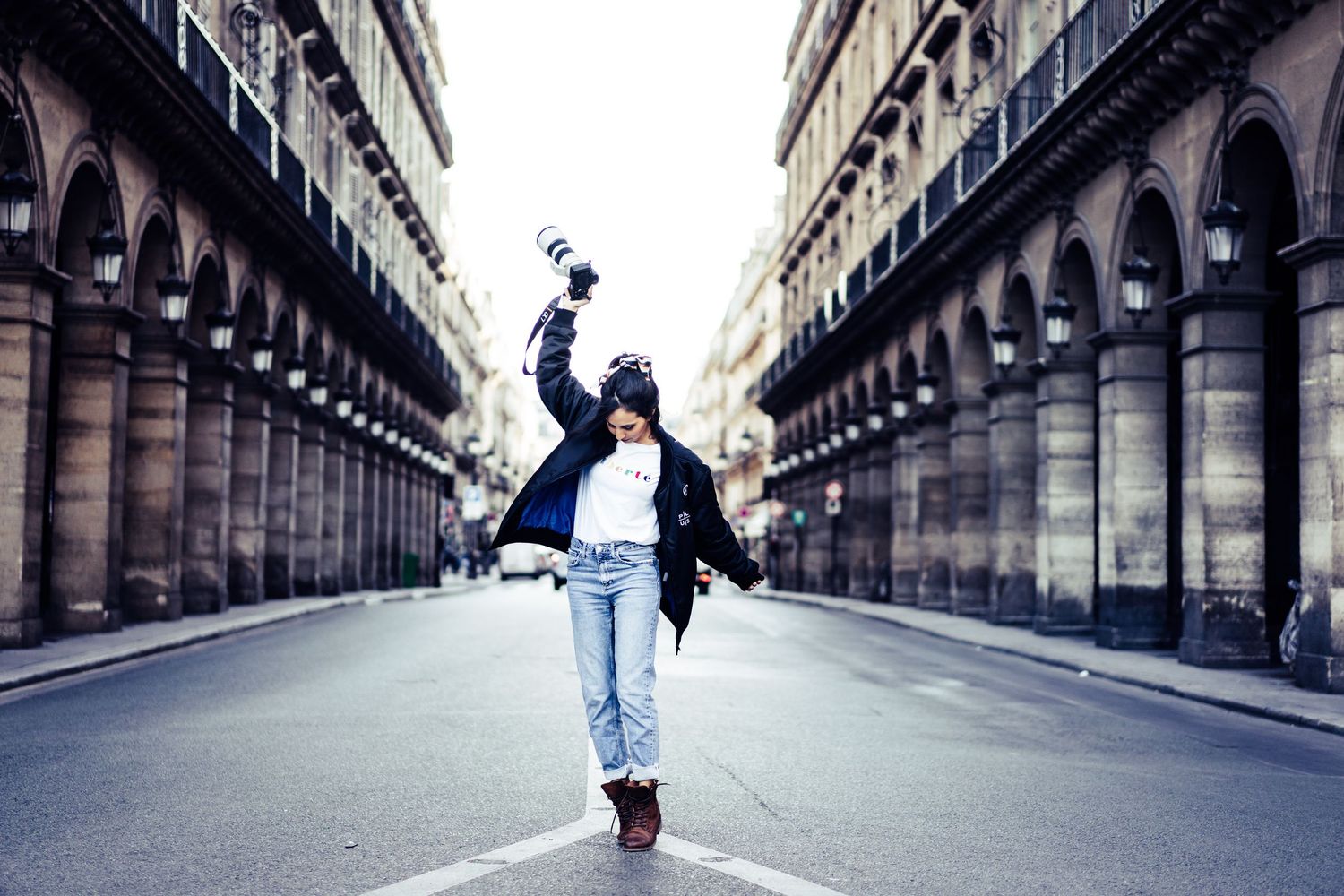 Person in casual winter outfit joyfully walking down an empty Parisian arcade street holding a camera.