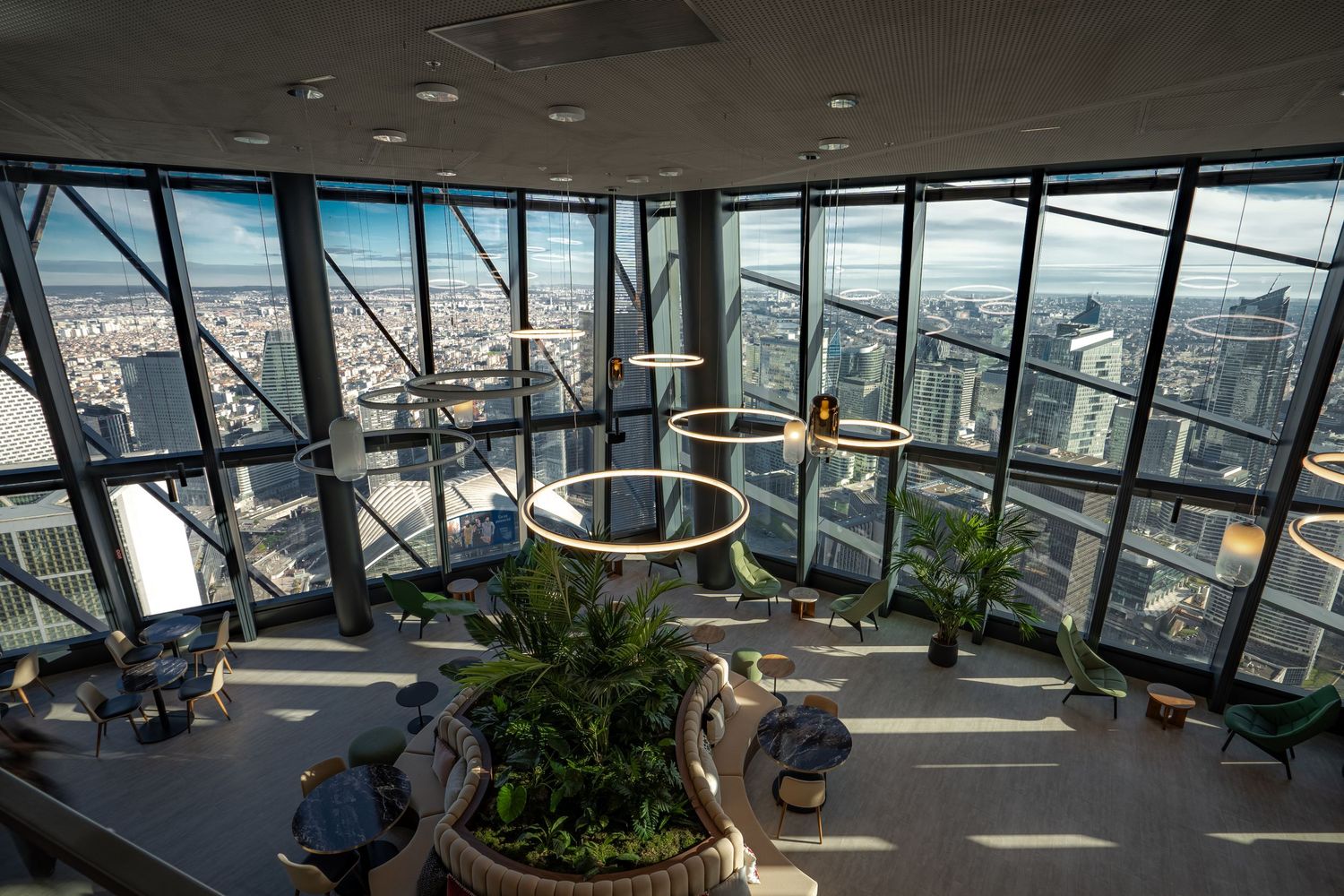 Stunning high-rise office lounge with panoramic city views, circular pendant lights, lush plants, and modern seating.