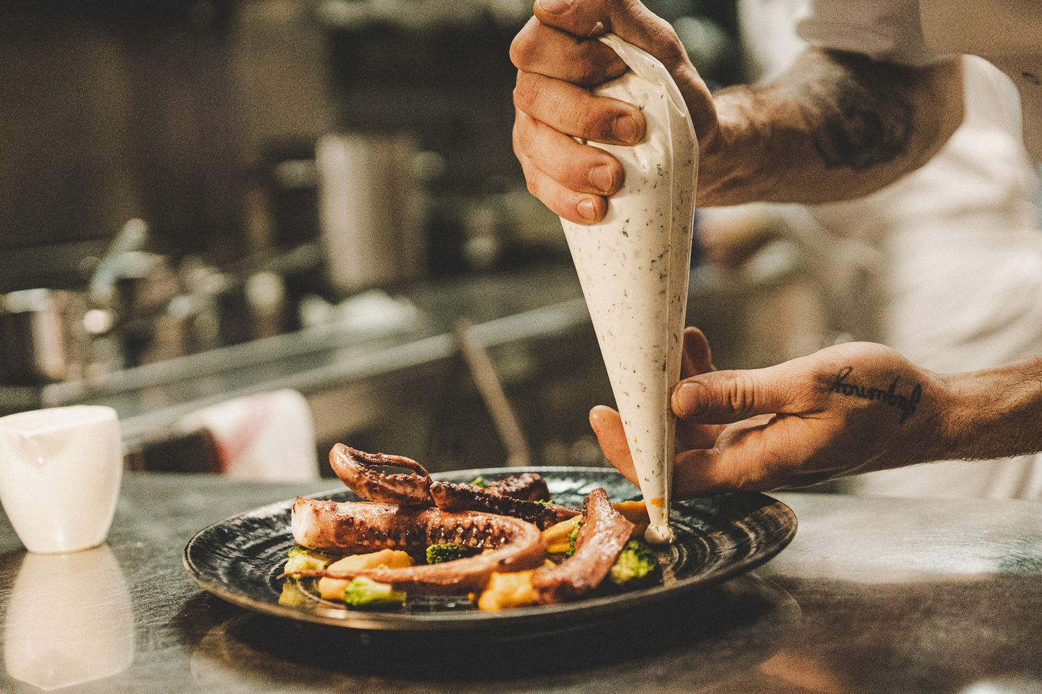 Tattooed chef piping sauce onto a gourmet octopus dish in a professional kitchen.