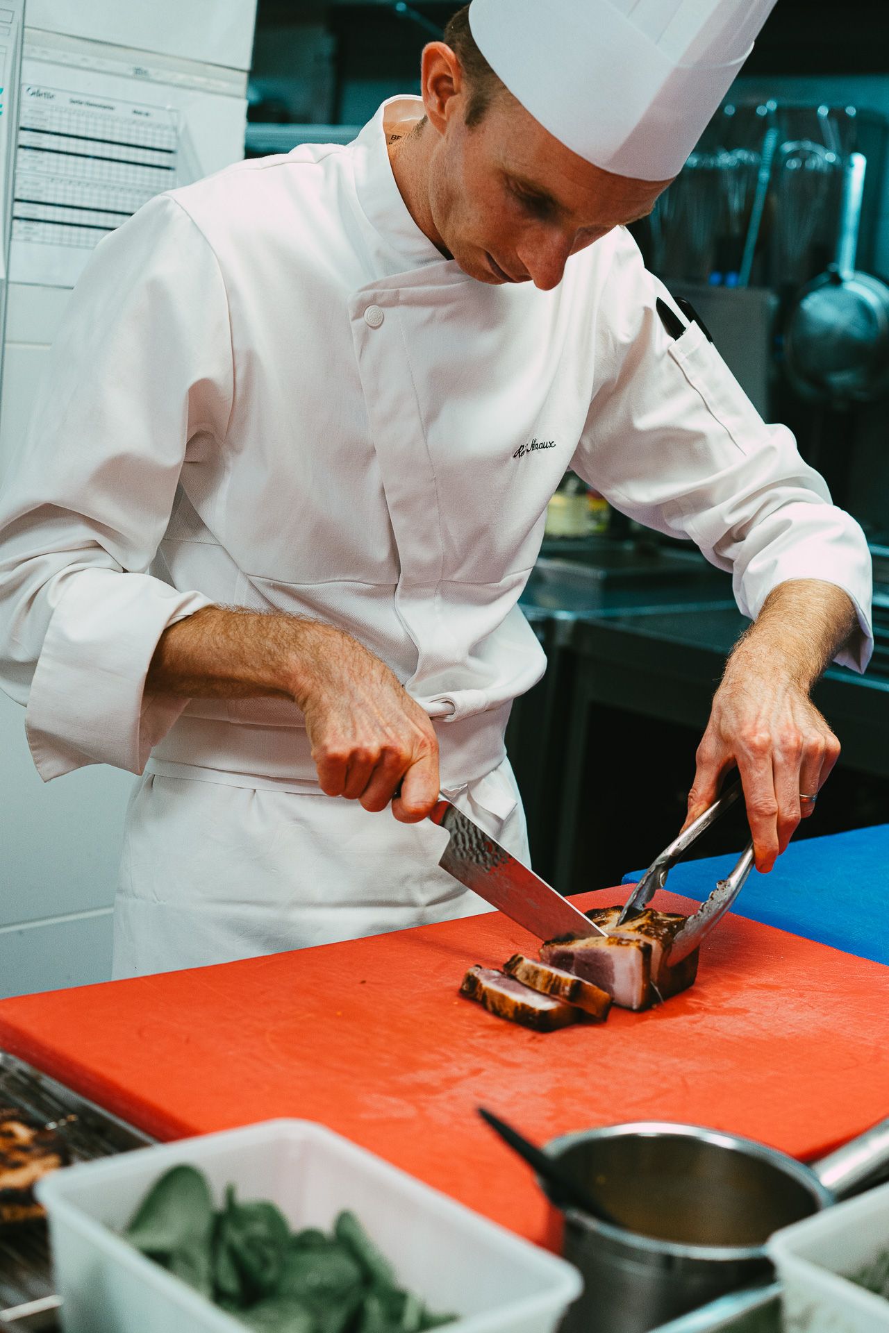 A professional chef in white uniform slices cooked meat on a red cutting board in a restaurant kitchen.