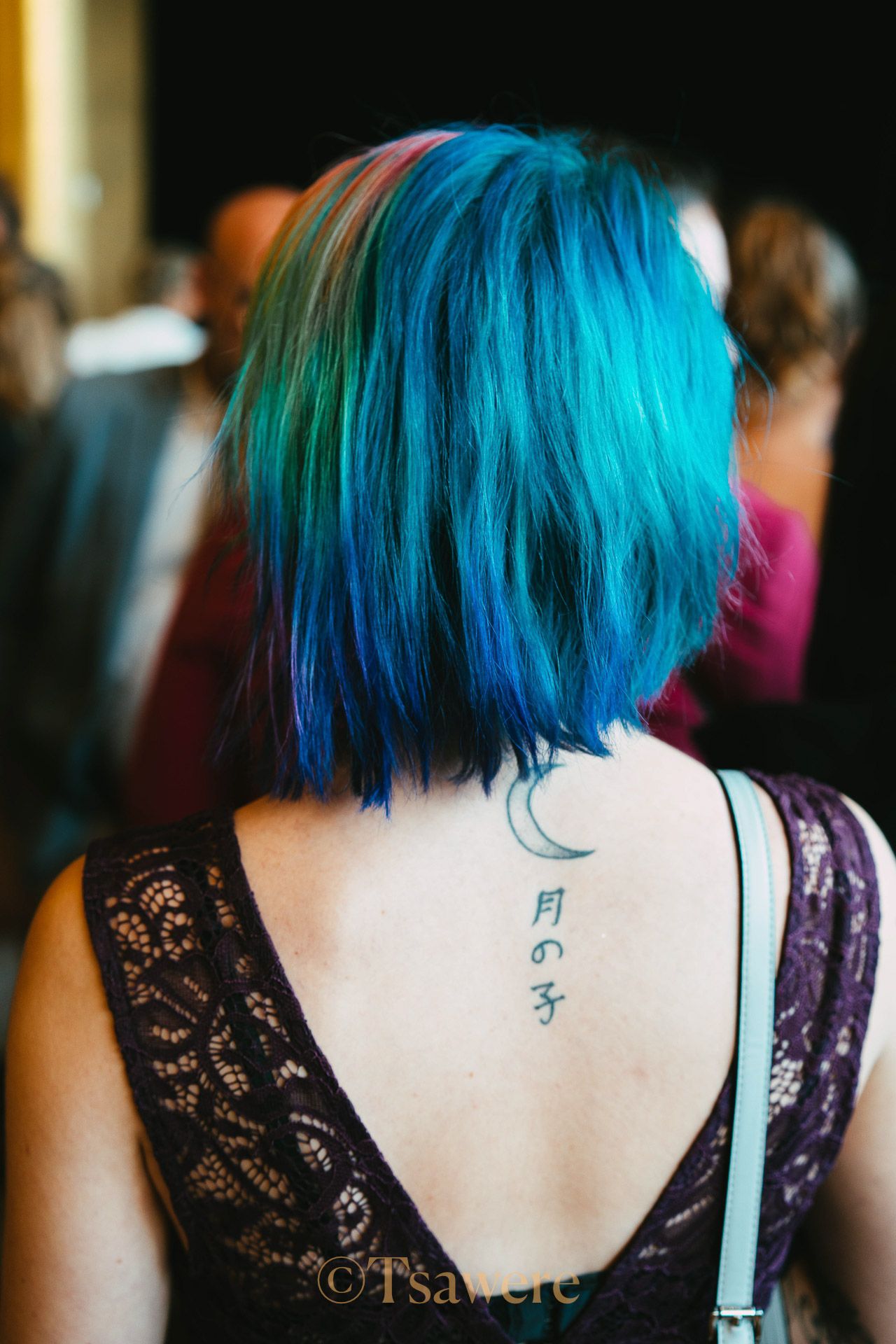 Woman with vibrant blue and green bob haircut and crescent moon tattoo on her back.