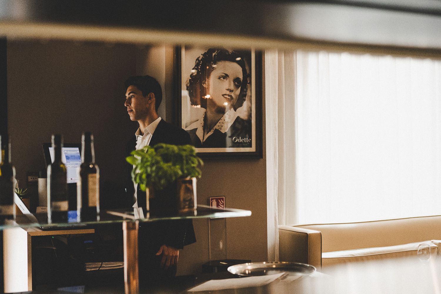 A man stands near a bar with wine bottles and a vintage portrait poster on the wall in a dimly lit restaurant.