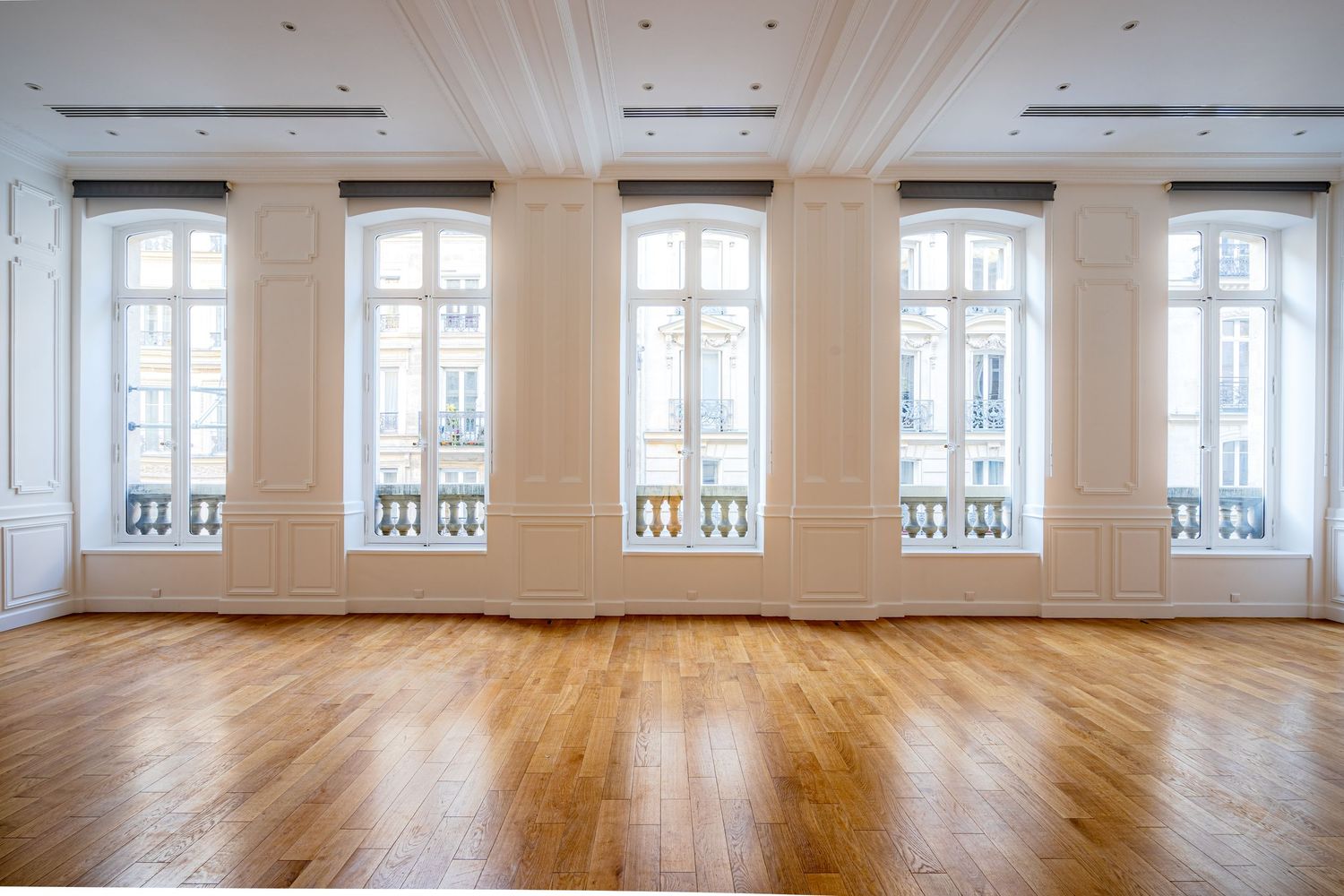 Empty grand Parisian room with ornate arched windows, decorative moldings, columns, and polished hardwood floors.