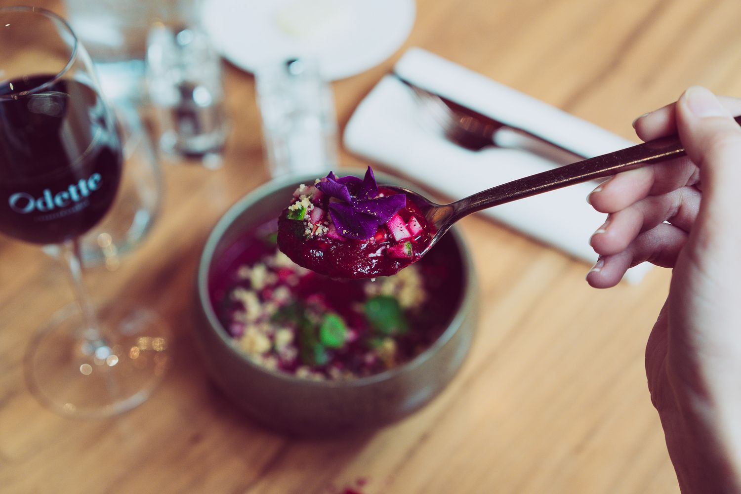 Hand holding a spoon with beet and quinoa soup at an Odette restaurant table setting.