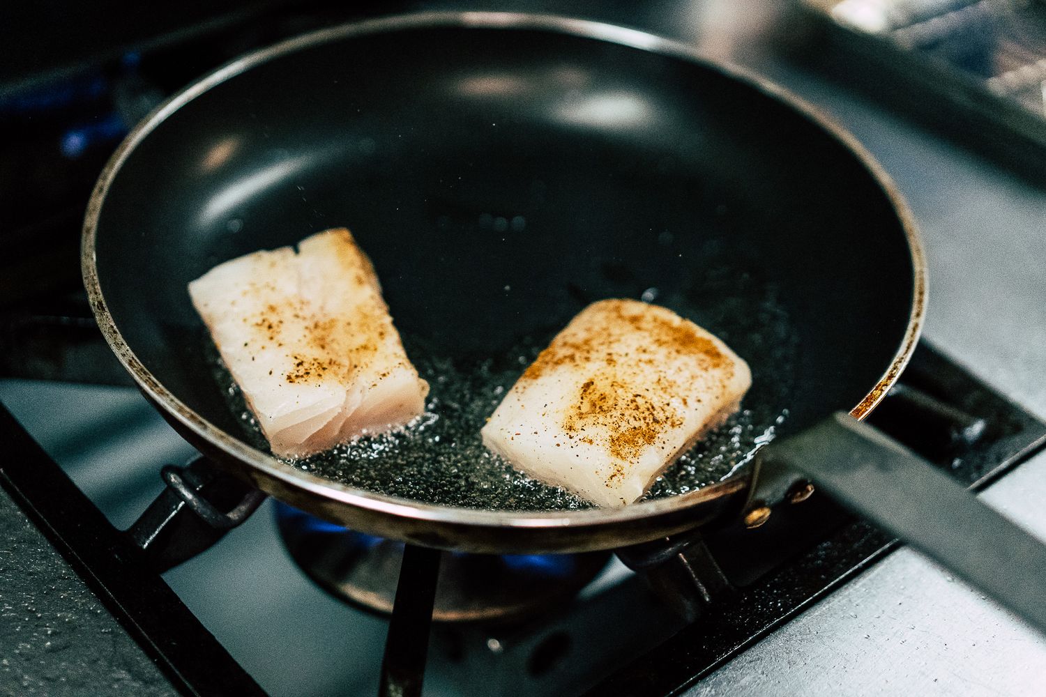 Two seasoned fish fillets sizzling in butter in a black non-stick frying pan on a gas stove.