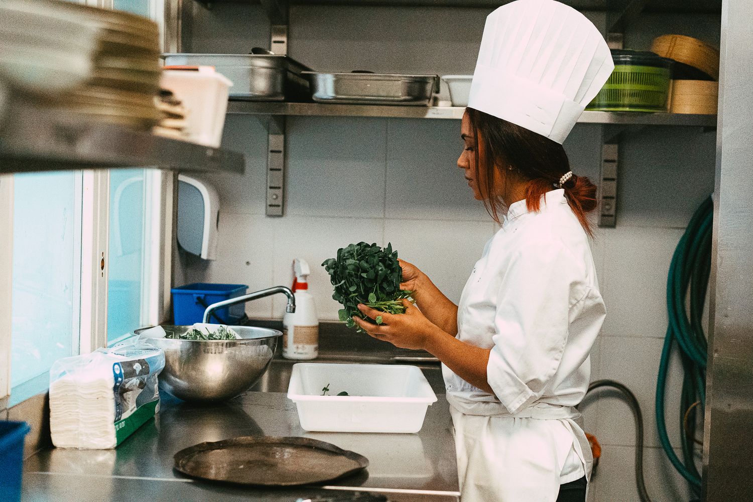 A female chef in white uniform and hat inspects fresh herbs at a stainless steel kitchen prep station.
