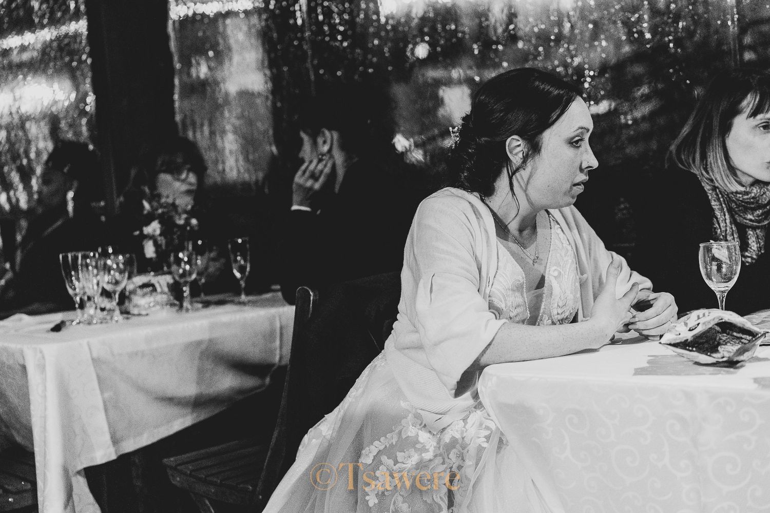 Bride seated at reception table with guests, candid black and white wedding photo.
