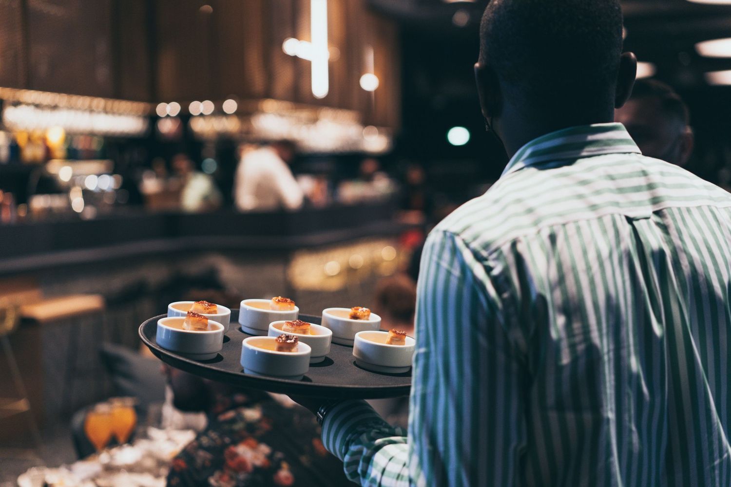 Waiter in striped shirt carrying a tray of small gourmet appetizers through a busy restaurant.