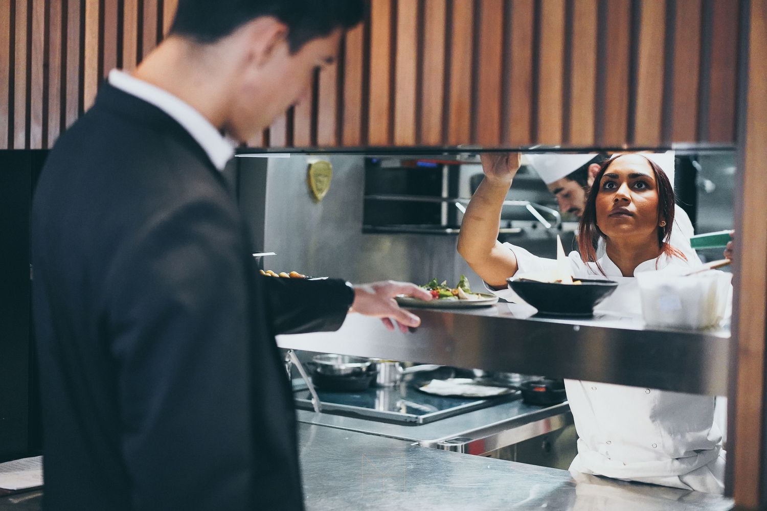 Chef passing plated dish through kitchen pass to suited waiter in modern restaurant kitchen.