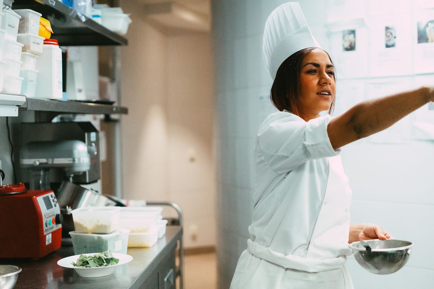Female chef in white uniform and hat reaching for ingredients in a professional restaurant kitchen.