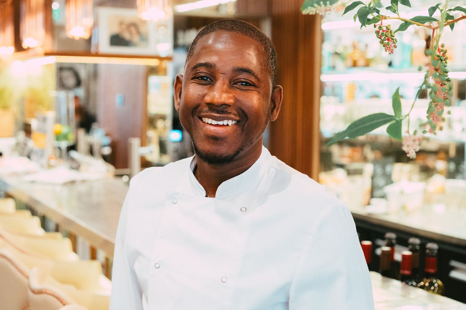 Smiling male chef in white uniform standing in a warmly lit upscale restaurant setting.