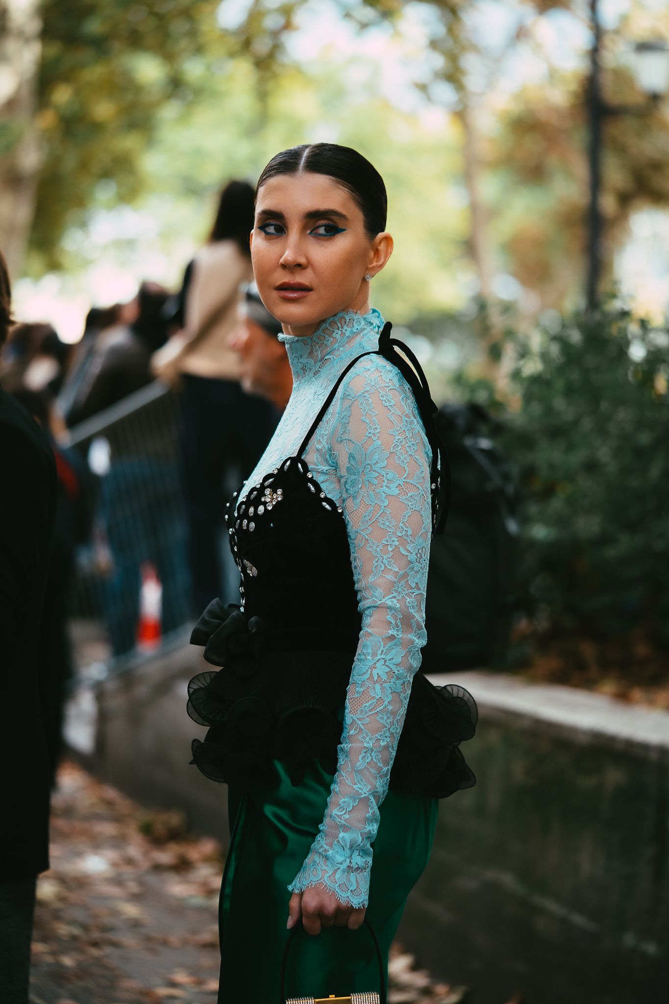 Woman in teal lace turtleneck under black ruffled dress posing at outdoor fashion week event.
