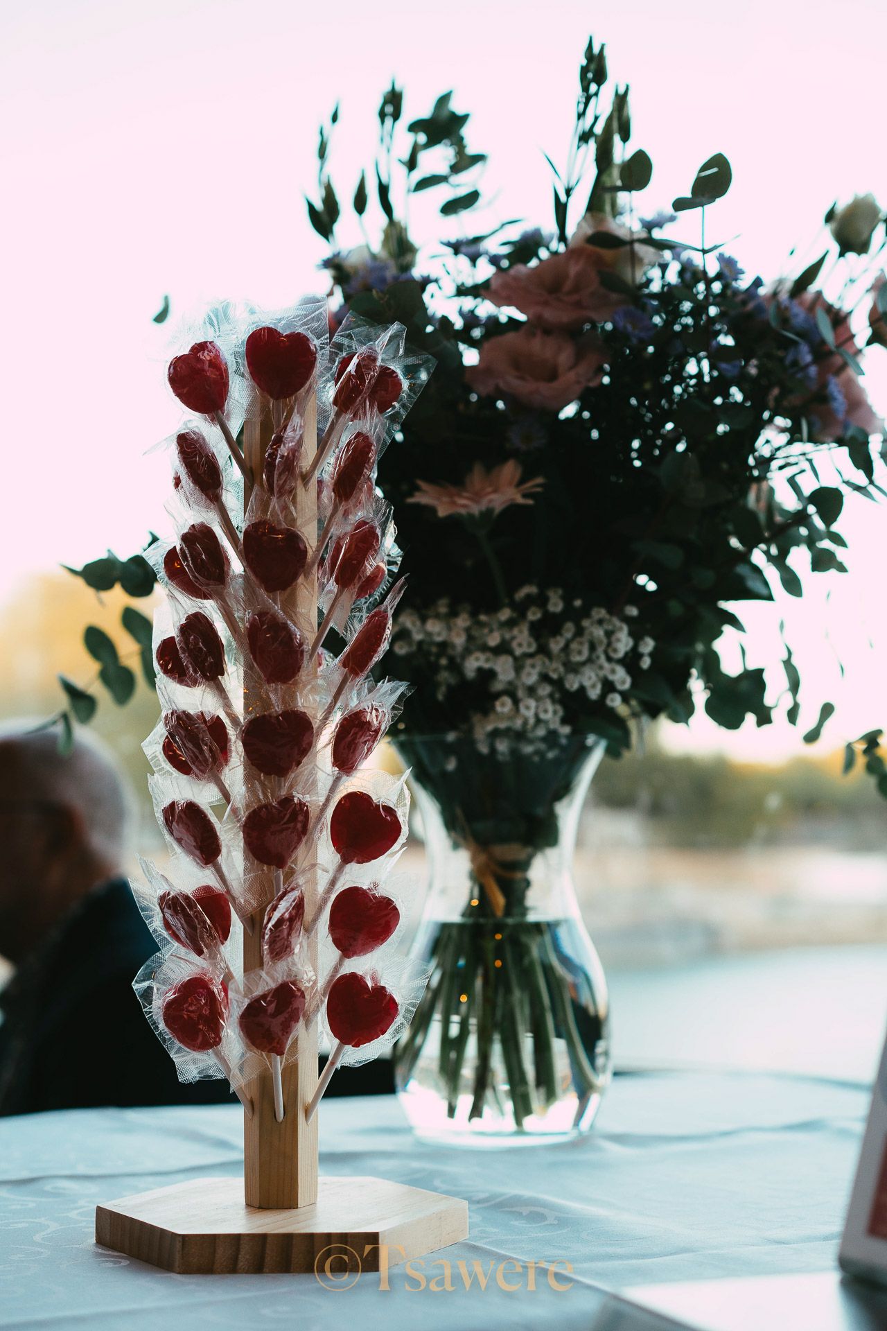 Heart-shaped lollipop wedding favor display beside floral bouquet centerpiece at reception table.