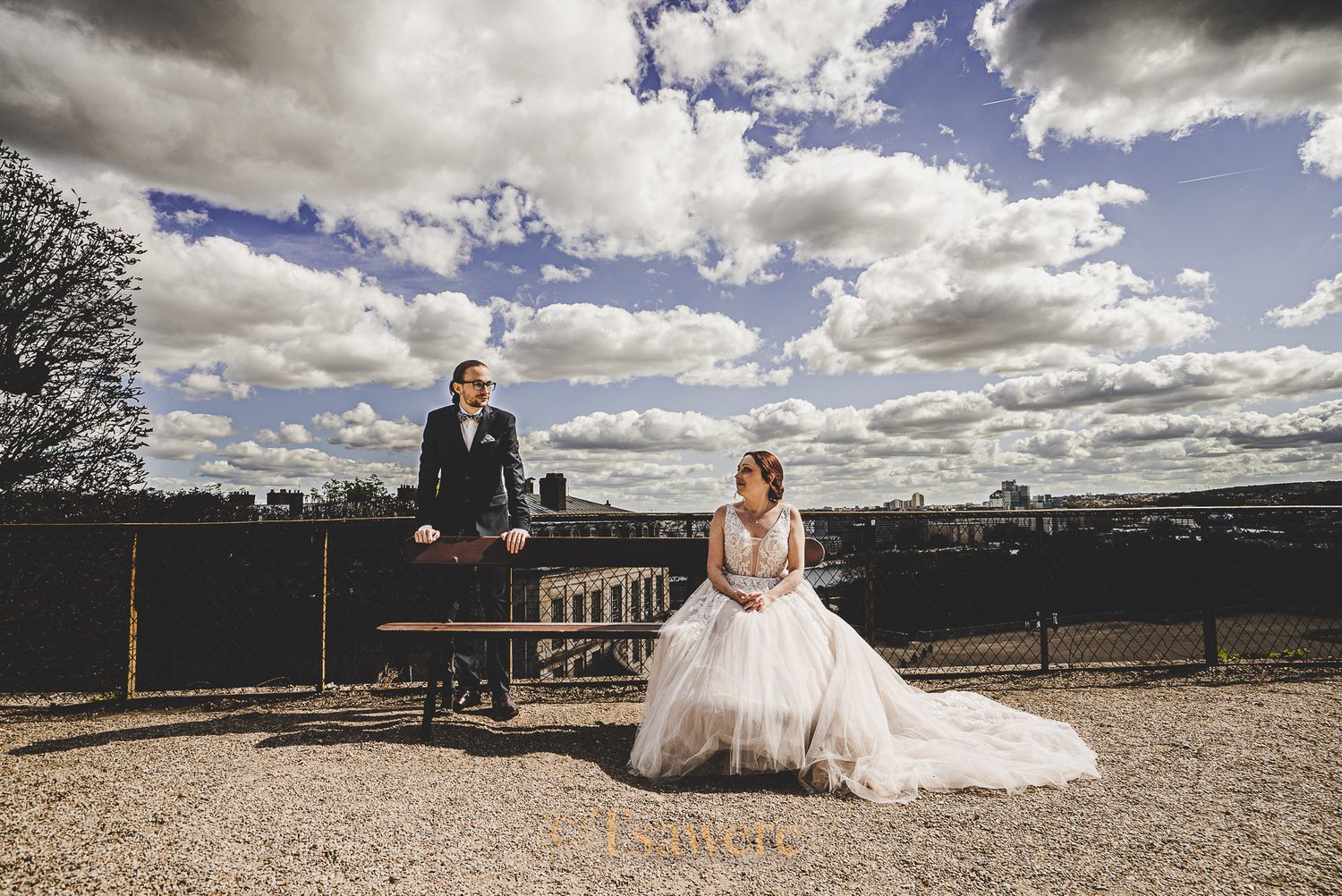 Bride in tulle gown and groom sitting apart on bench with dramatic cloudy sky backdrop.