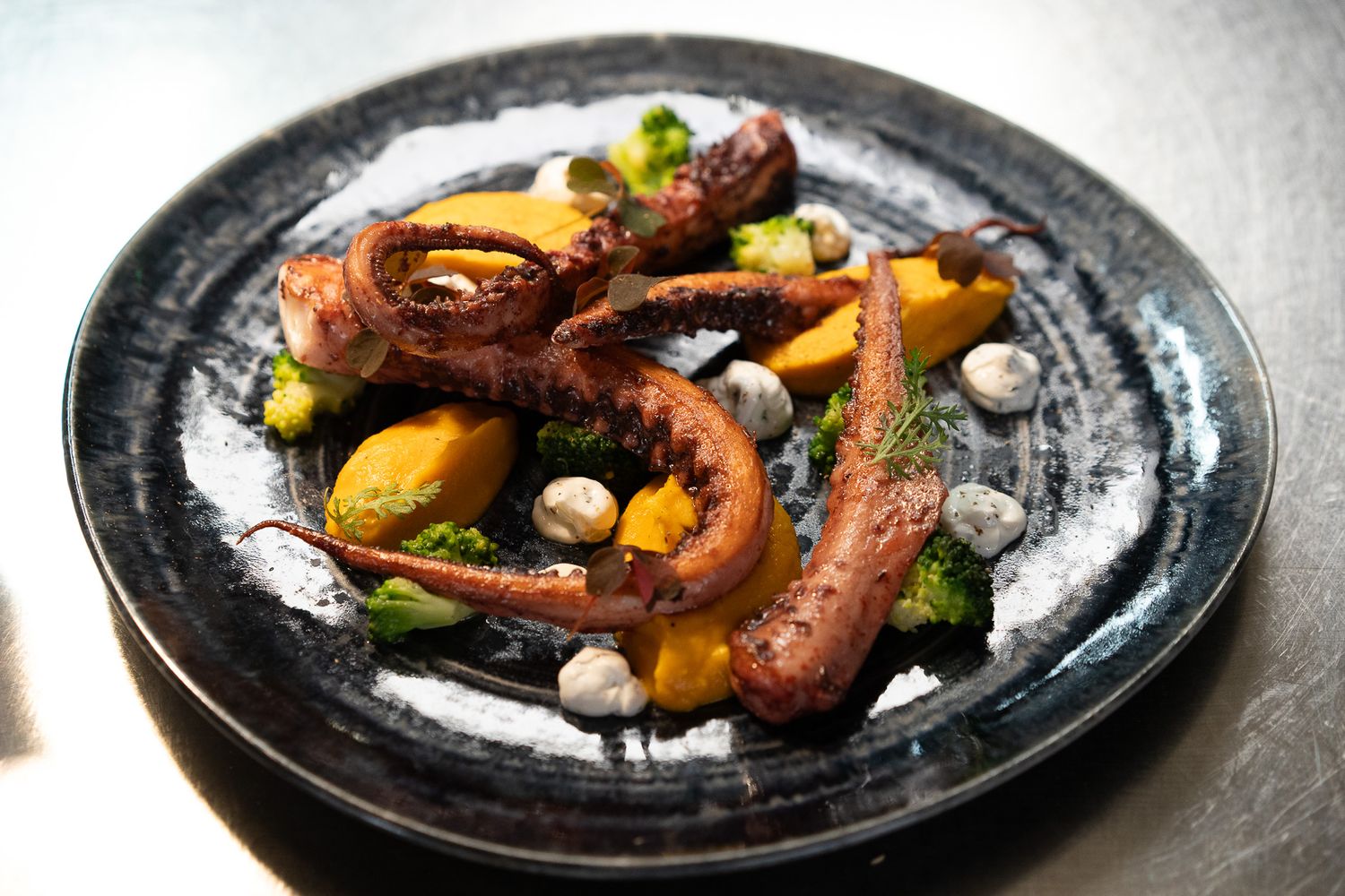 Artistic plating of grilled octopus with vegetables on a dark ceramic plate.