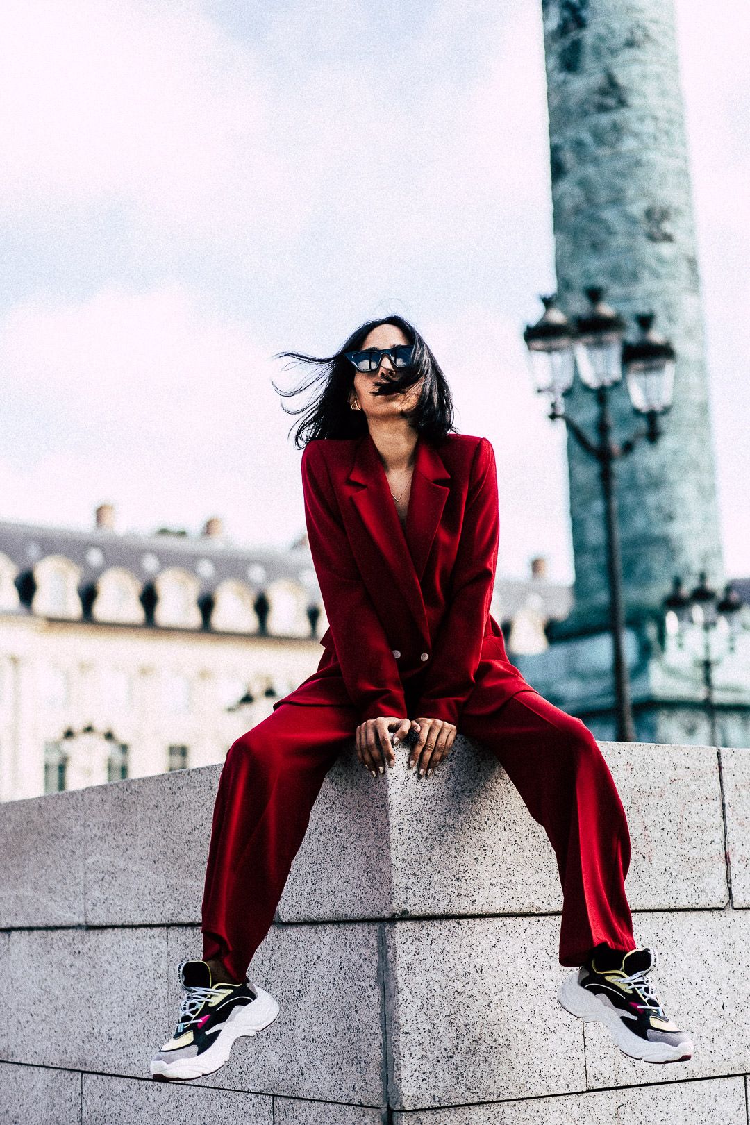 Stylish woman in red pantsuit and sneakers sitting on stone ledge in Paris square.