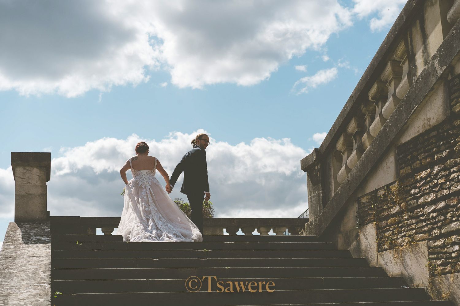 Bride and groom holding hands while ascending grand stone staircase under dramatic cloudy sky.