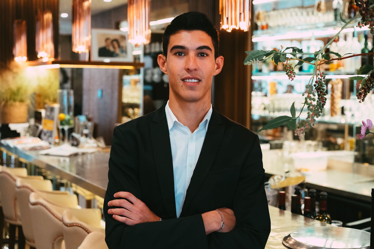 A young man in a black blazer stands with arms crossed in an elegant restaurant bar.