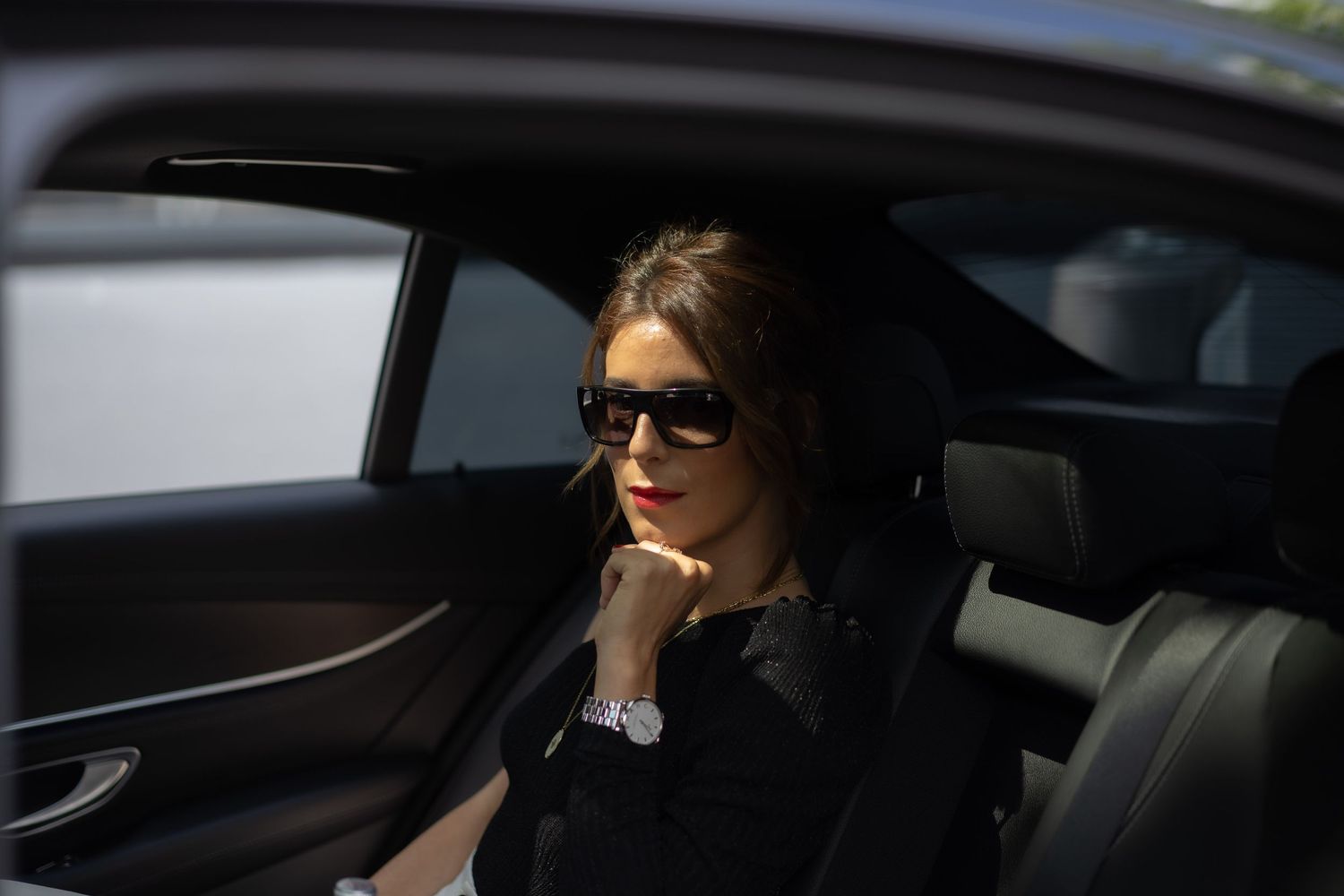 Stylish woman in black outfit and sunglasses sitting in luxury car backseat.