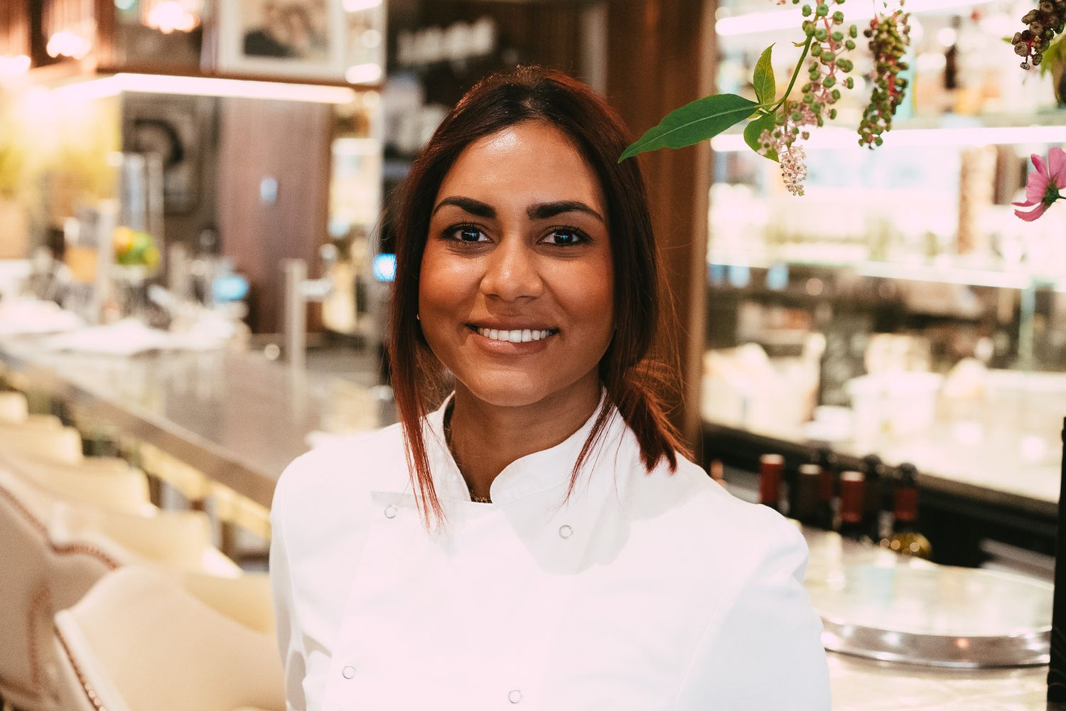 A smiling female chef in a white uniform poses in a modern restaurant kitchen.