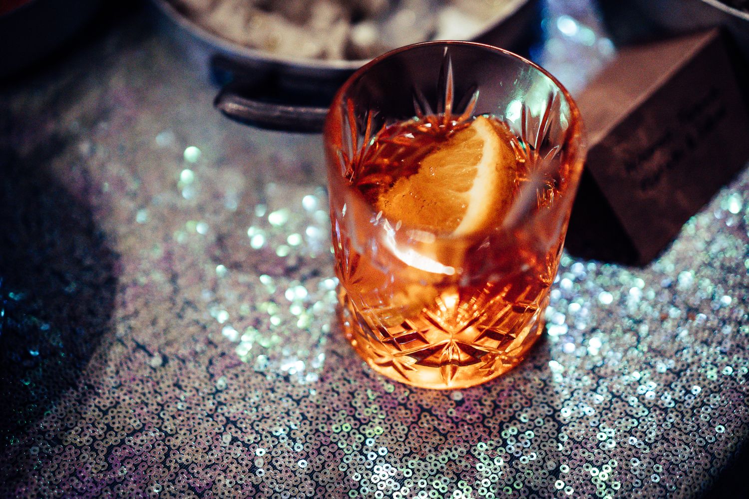 A crystal rocks glass with an orange peel garnish cocktail on a silver sequin surface.