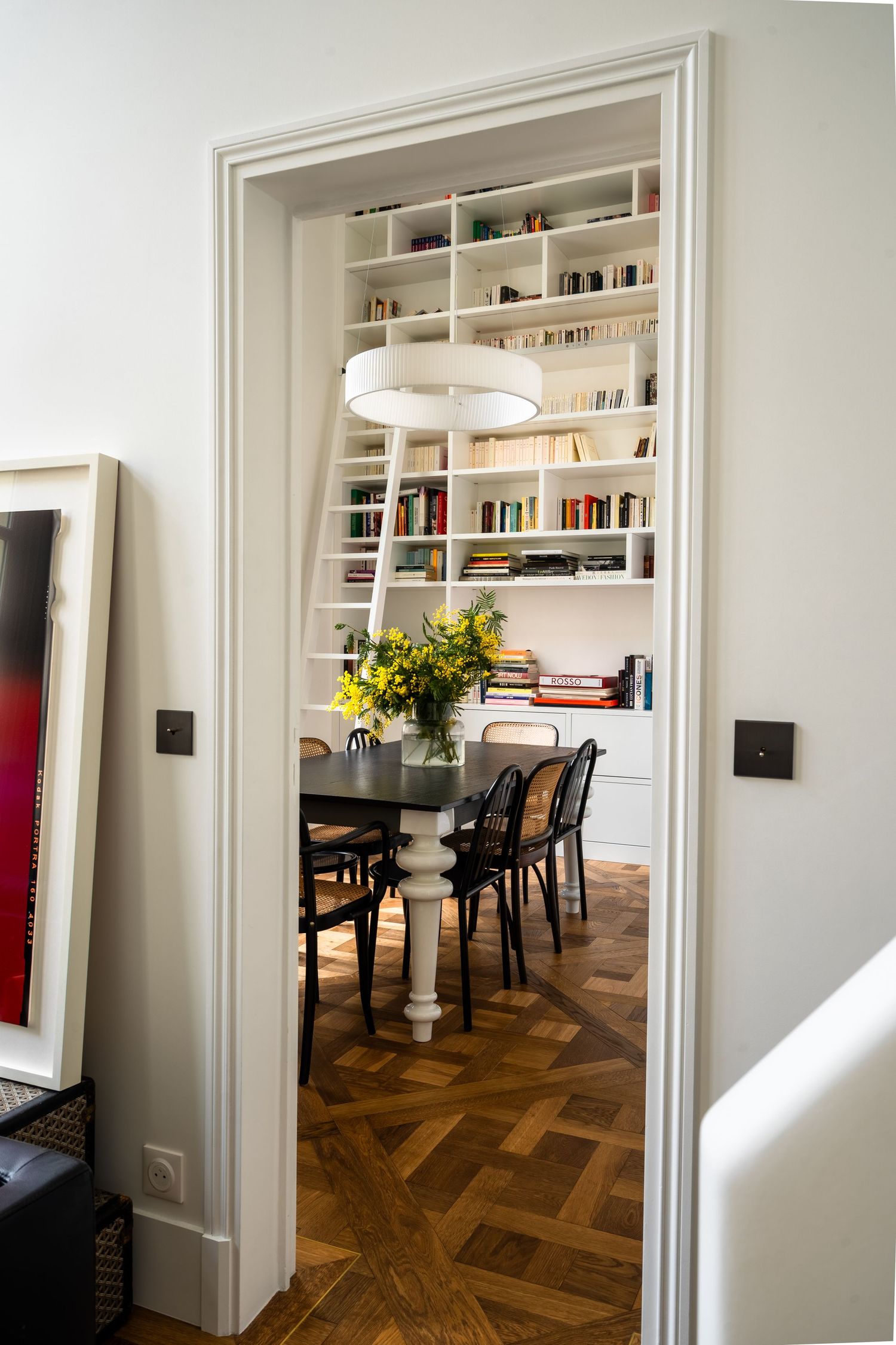 Doorway view into a sophisticated dining room featuring built-in white bookshelves, black dining table, and herringbone parquet floor.