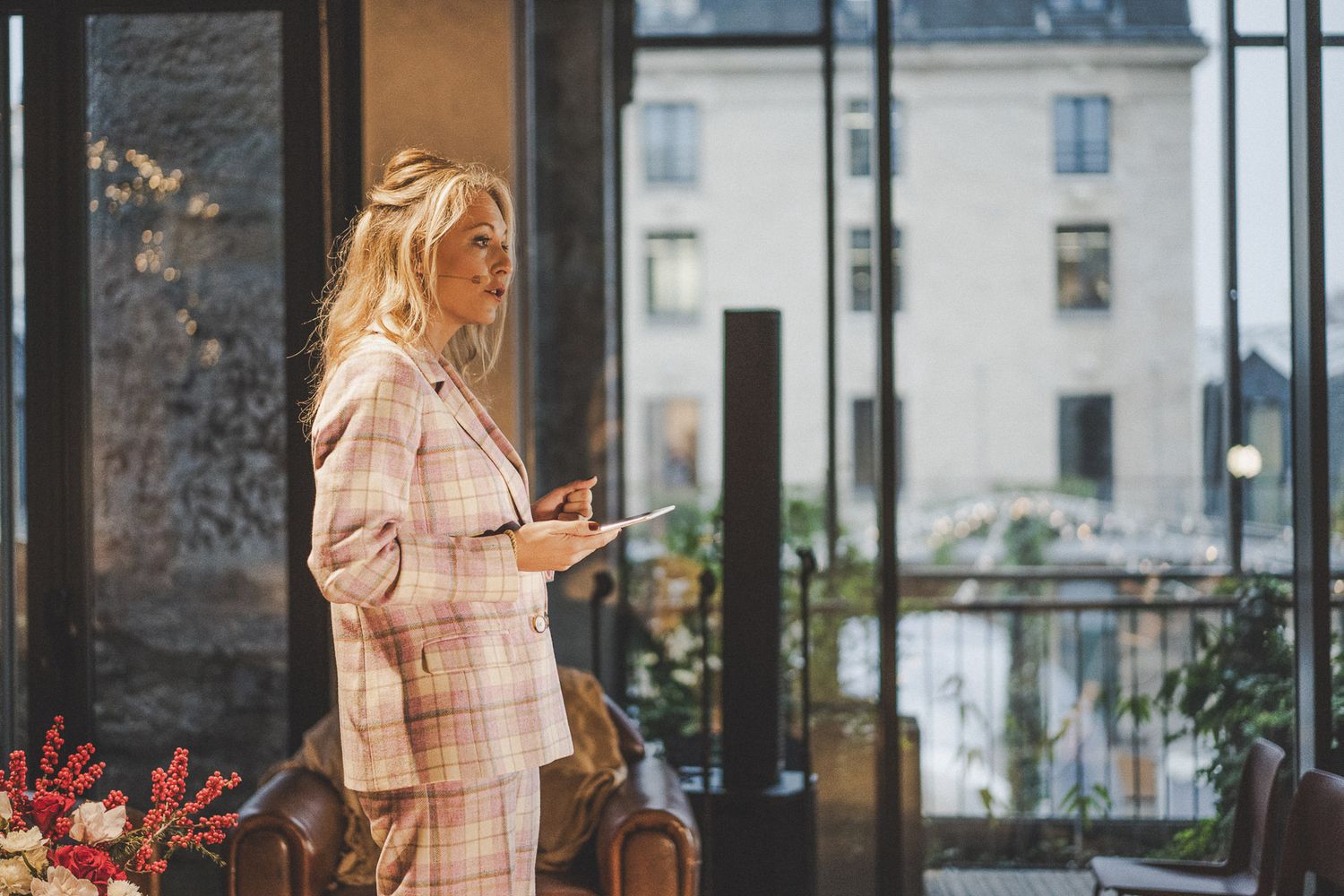Elegant blonde woman in pink plaid suit holding tablet beside large windows in stylish interior.