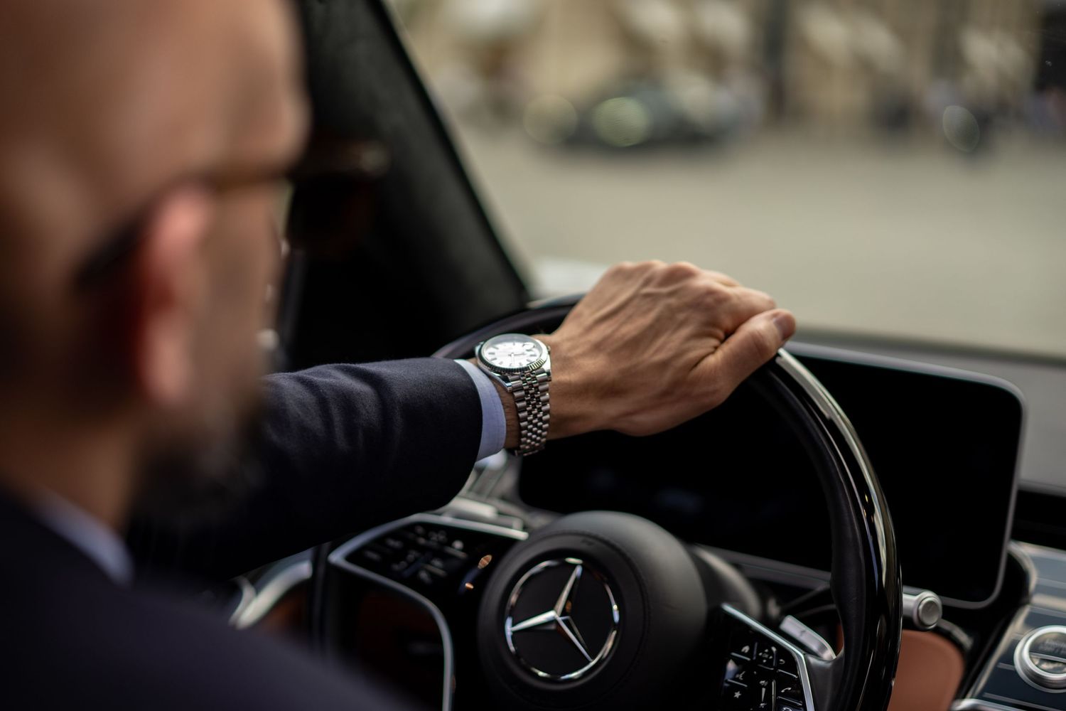 Close-up of man's hand on Mercedes-Benz steering wheel wearing a luxury watch in suit.