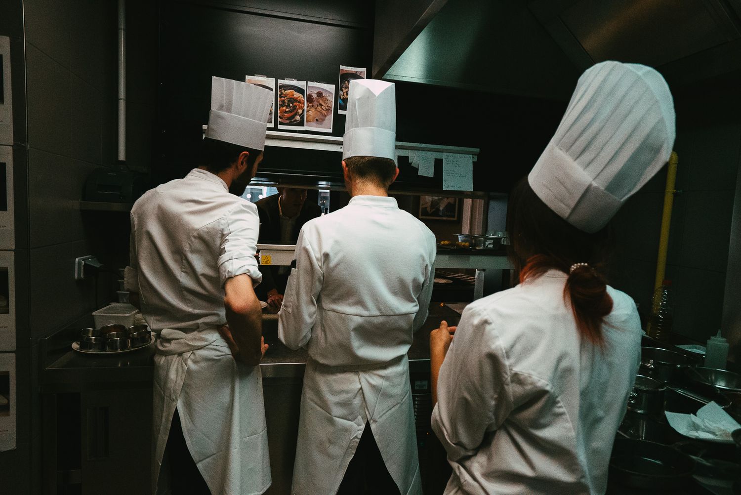 Three chefs in white uniforms reviewing recipes together at a commercial kitchen workstation.
