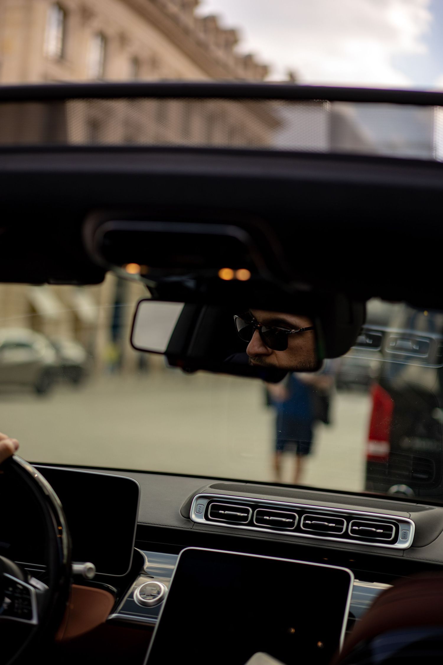 Driver in sunglasses reflected in rearview mirror of luxury Mercedes interior on city street.