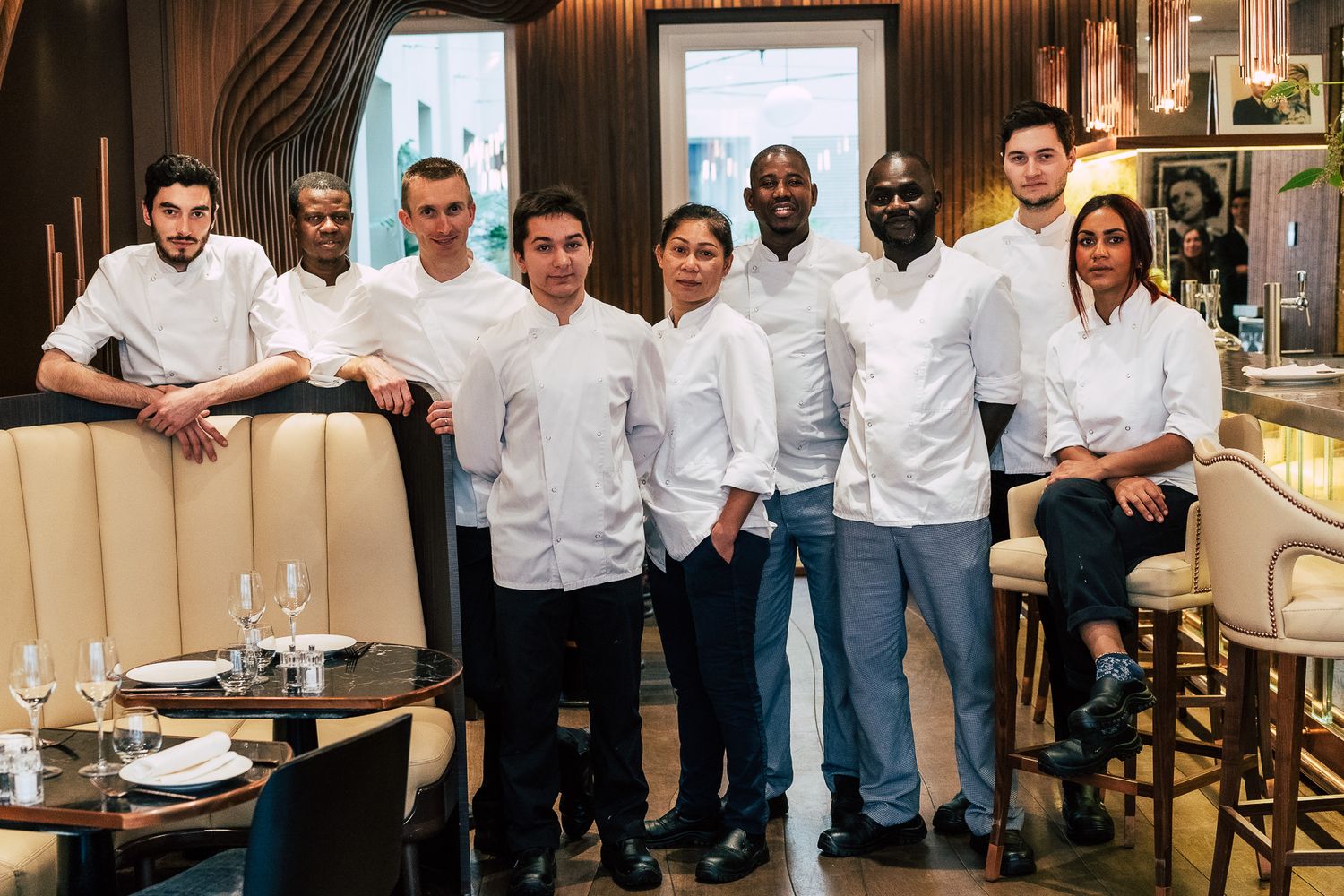 Professional culinary team of eight chefs in white uniforms posing together in an elegant restaurant dining room.