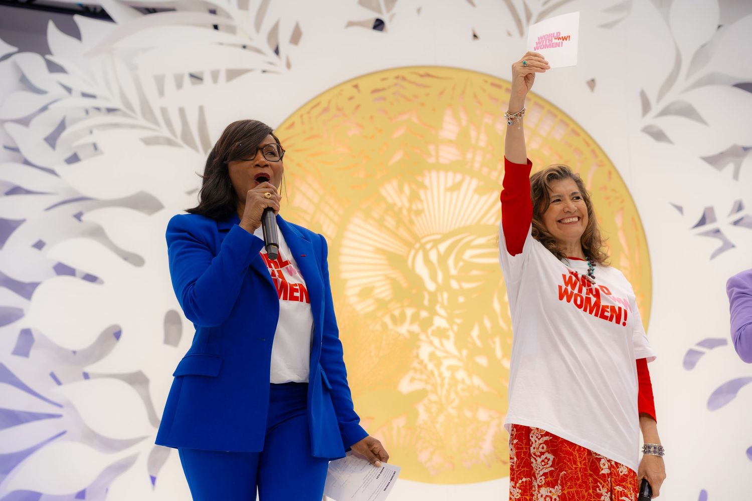 Two women on stage at a women empowerment event, one in blue suit, one in white shirt raising a paper.