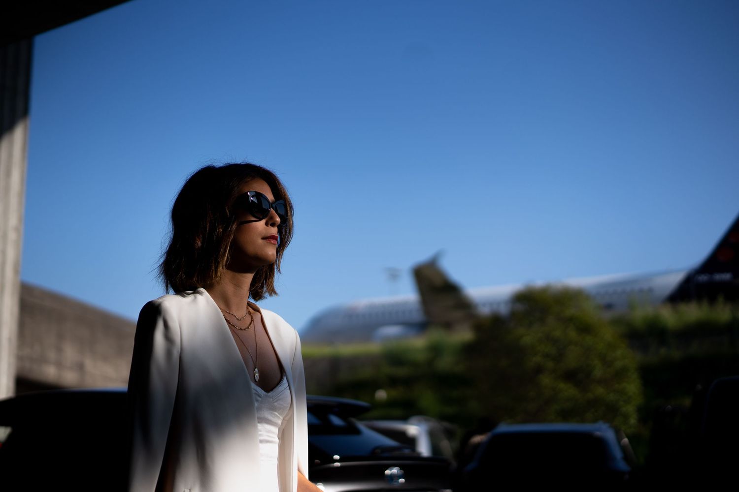 Stylish woman in white blazer wearing sunglasses outdoors on a sunny day near an airport terminal.