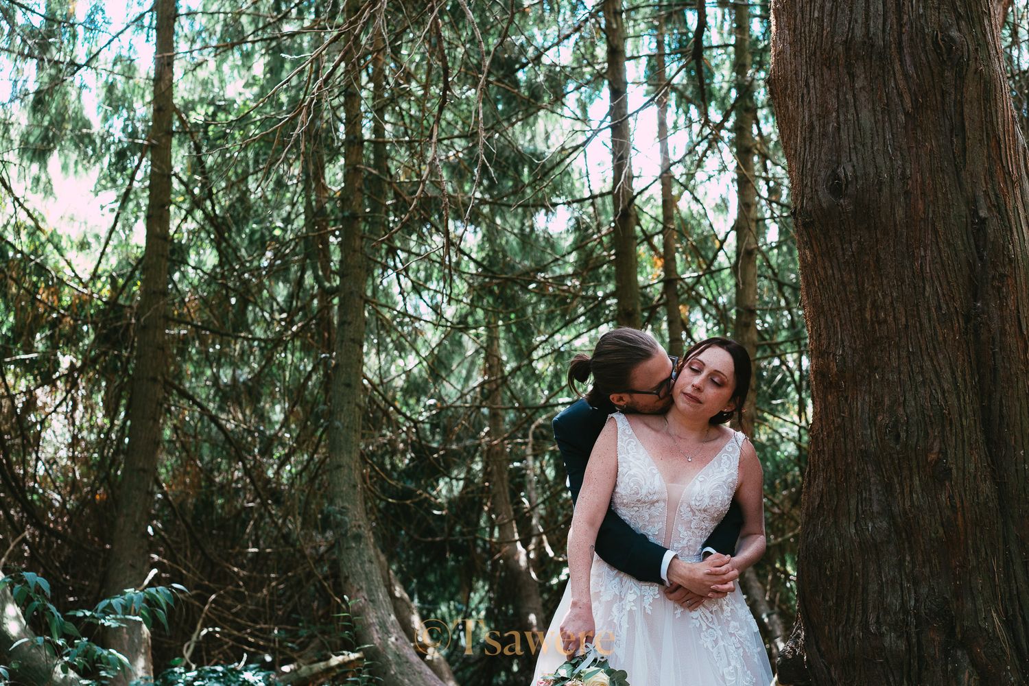 Groom kissing bride on cheek in a lush forest setting during wedding photo session.