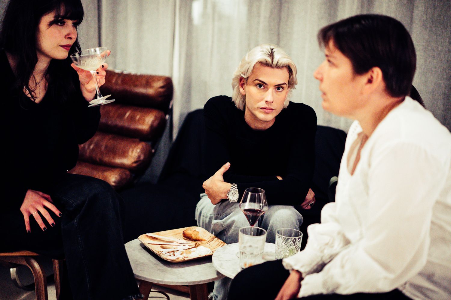 Three people socializing with drinks at a casual indoor gathering, seated around a coffee table.