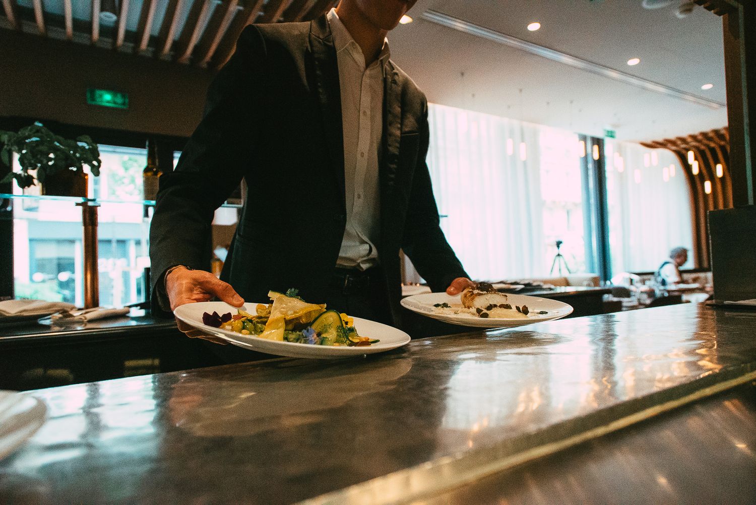 Restaurant server in black suit presenting two elegantly plated gourmet dishes on a metal counter.