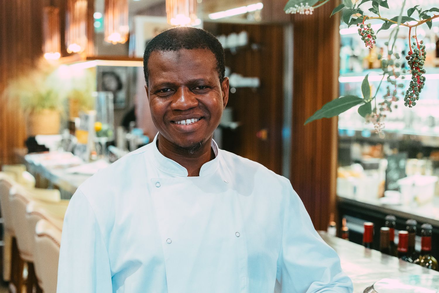 Cheerful male chef in light blue uniform smiling in a warmly lit fine dining restaurant.