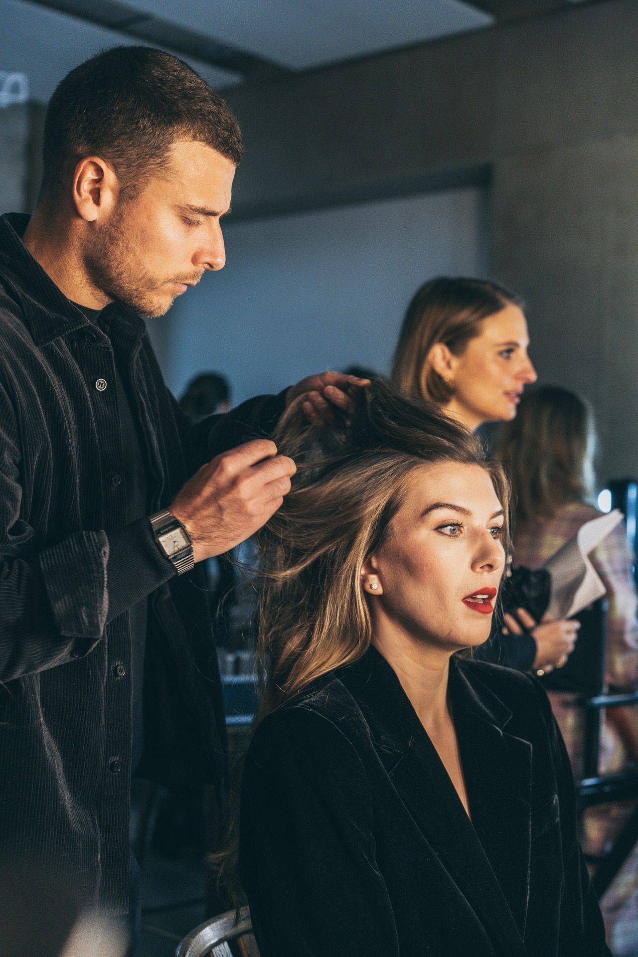 Male hairstylist styling a young brunette woman's hair backstage at a professional fashion event.