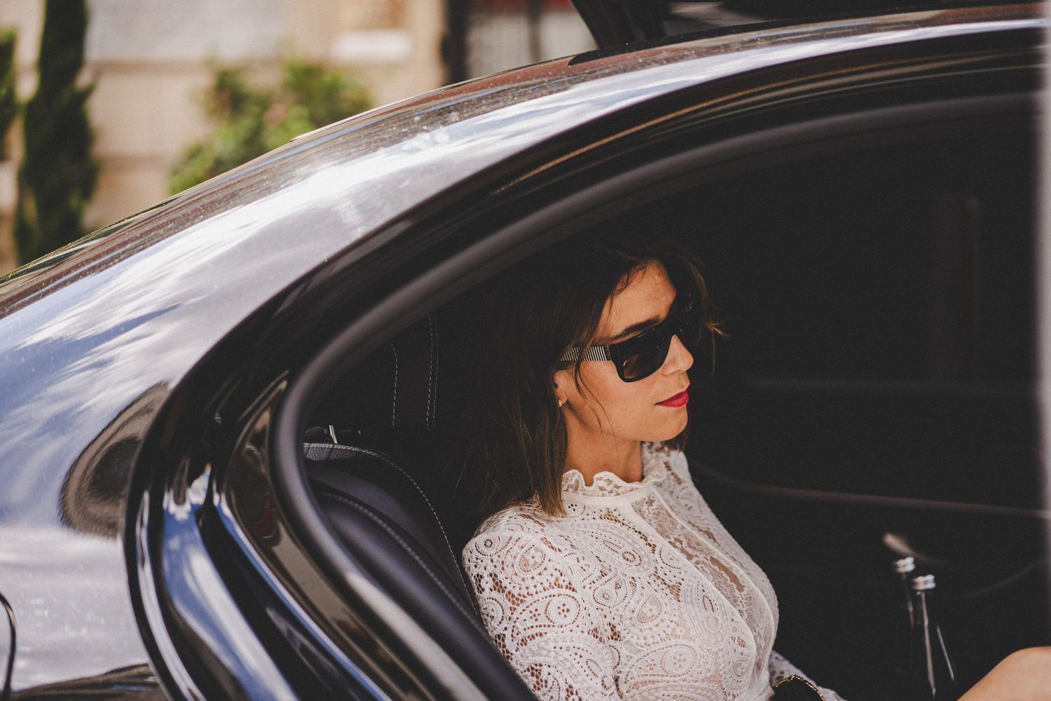 Sophisticated woman wearing sunglasses and white lace dress sitting in backseat of luxury vehicle.