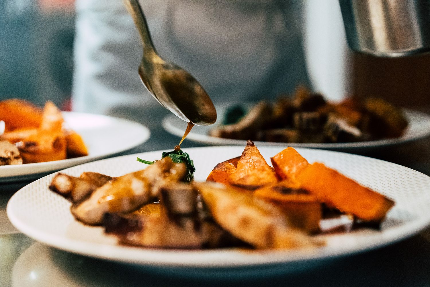 Chef drizzling green herb sauce over a plated dish with roasted vegetables and meat.