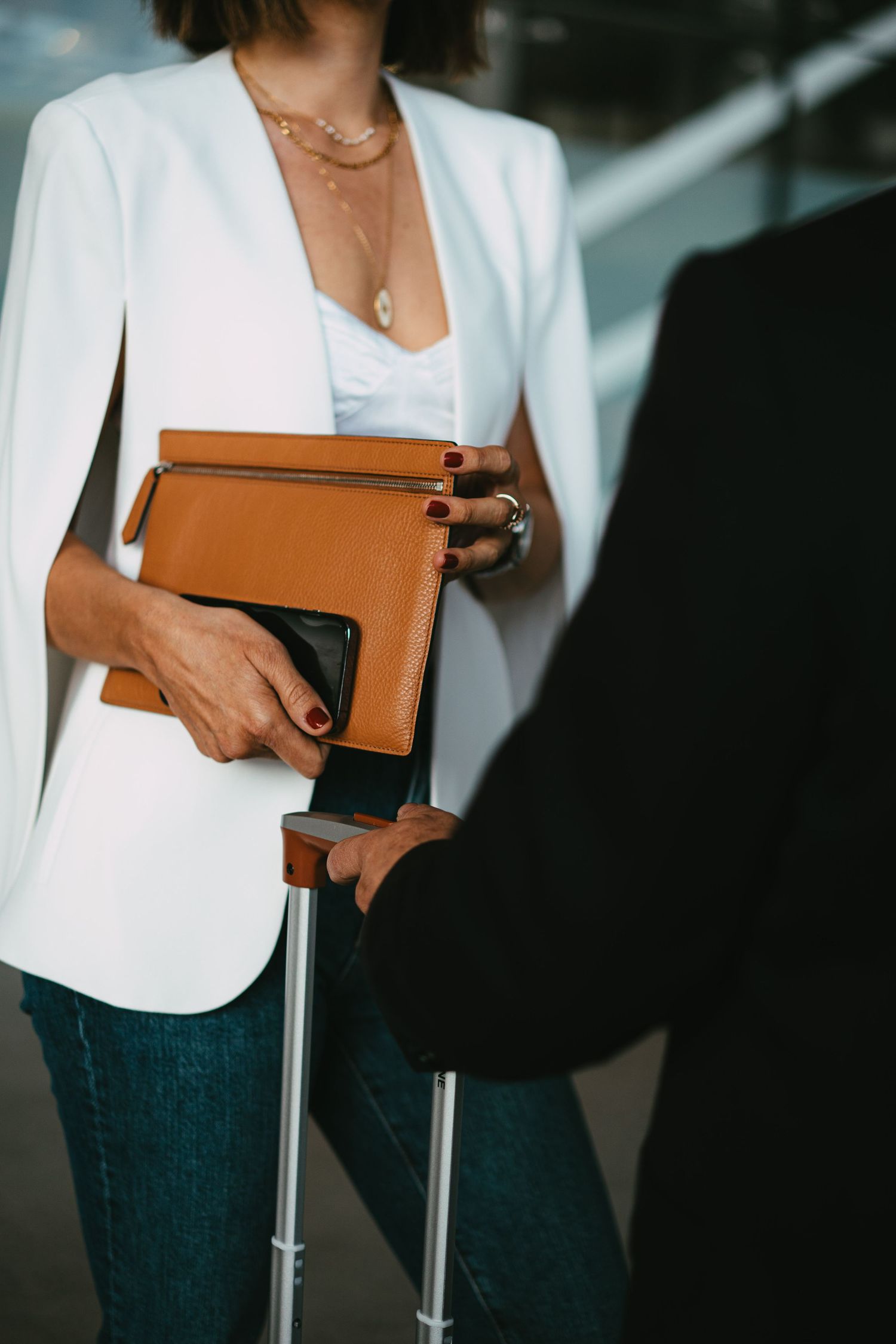Close-up of woman in white blazer holding brown leather clutch and passport near luggage at airport.