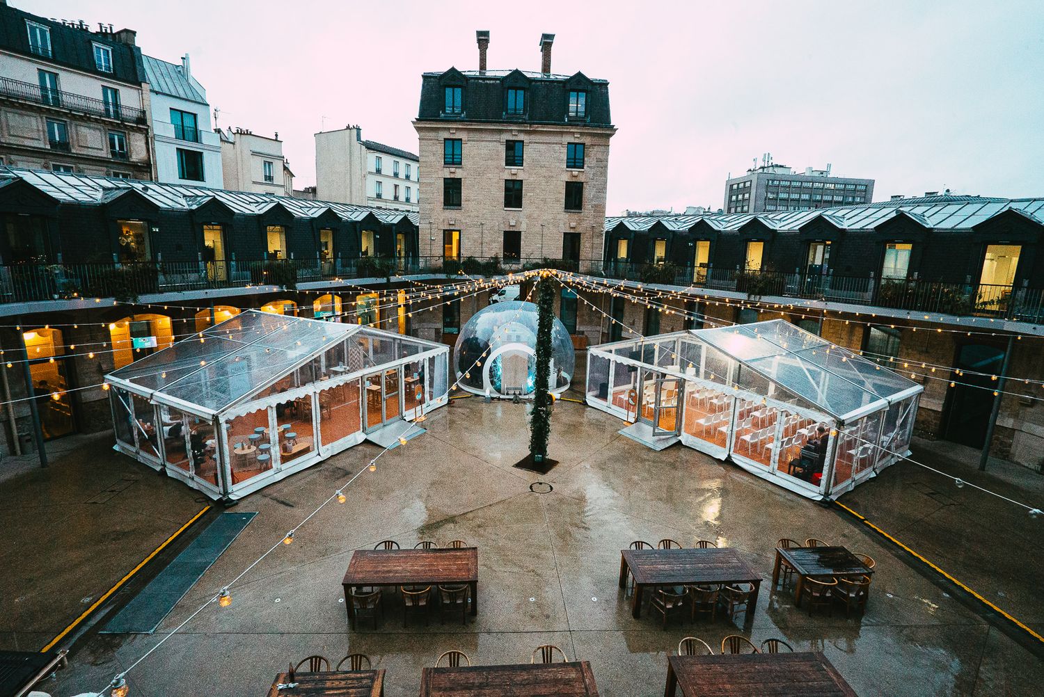 Aerial view of a Parisian courtyard with glass bubble dining tents and string lights at dusk.