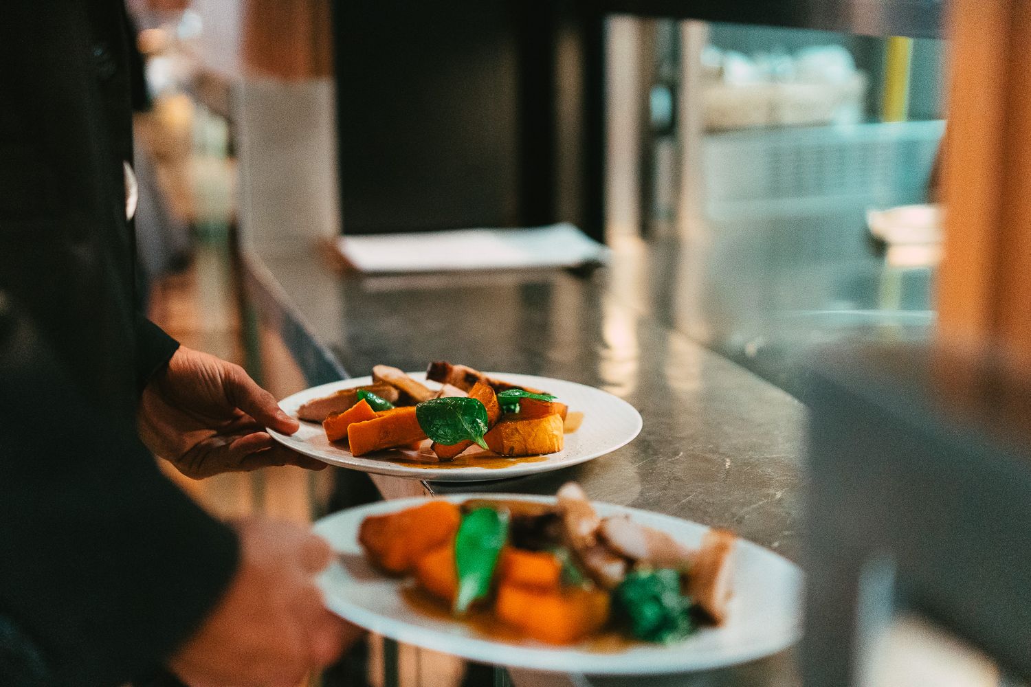 Server carrying two beautifully plated dishes with roasted vegetables through a restaurant.